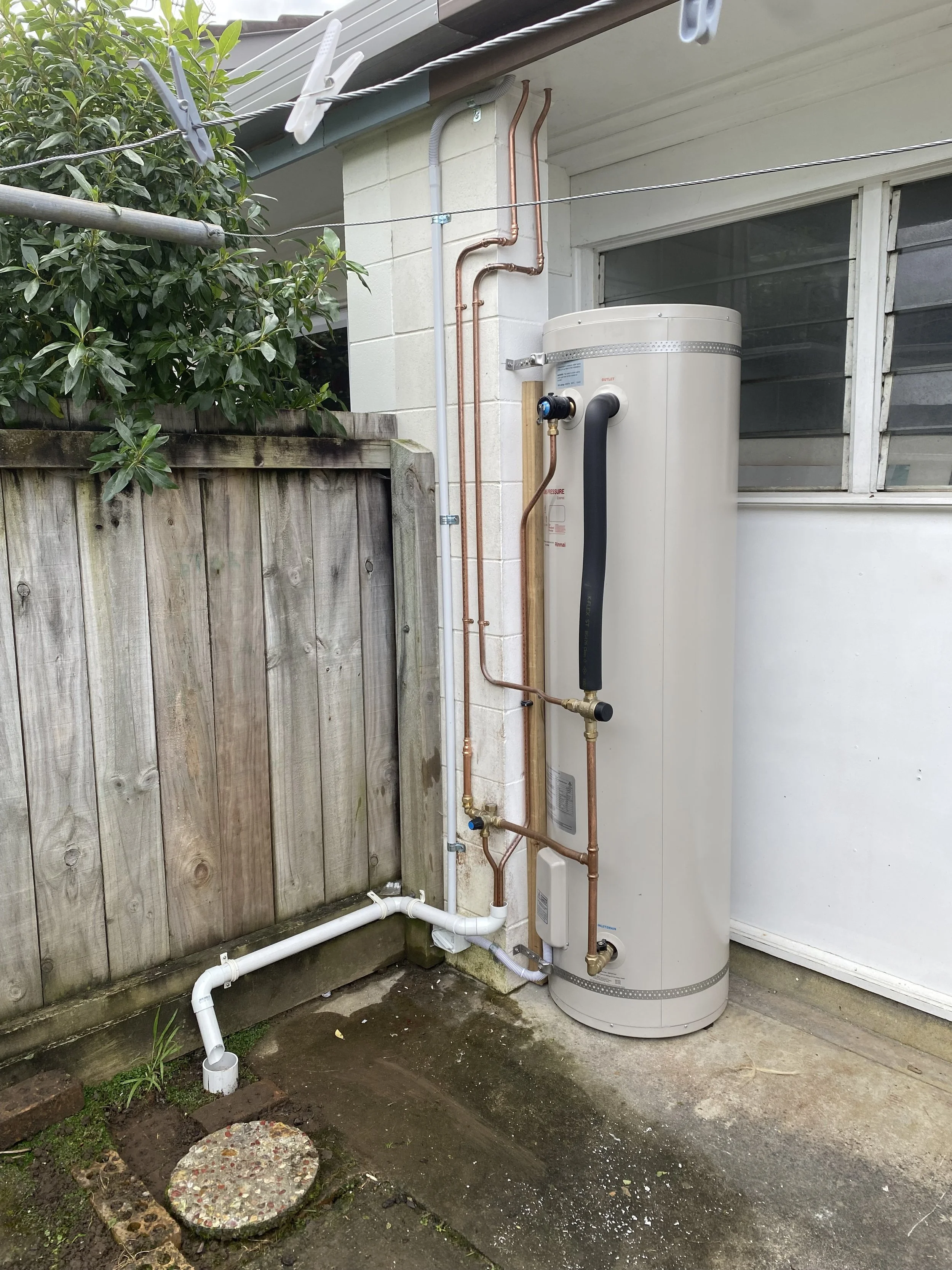 An outdoor scene showing a water heater, pipes and plumbing on the side of a house, with a wooden fence, a bush, and a concrete floor.