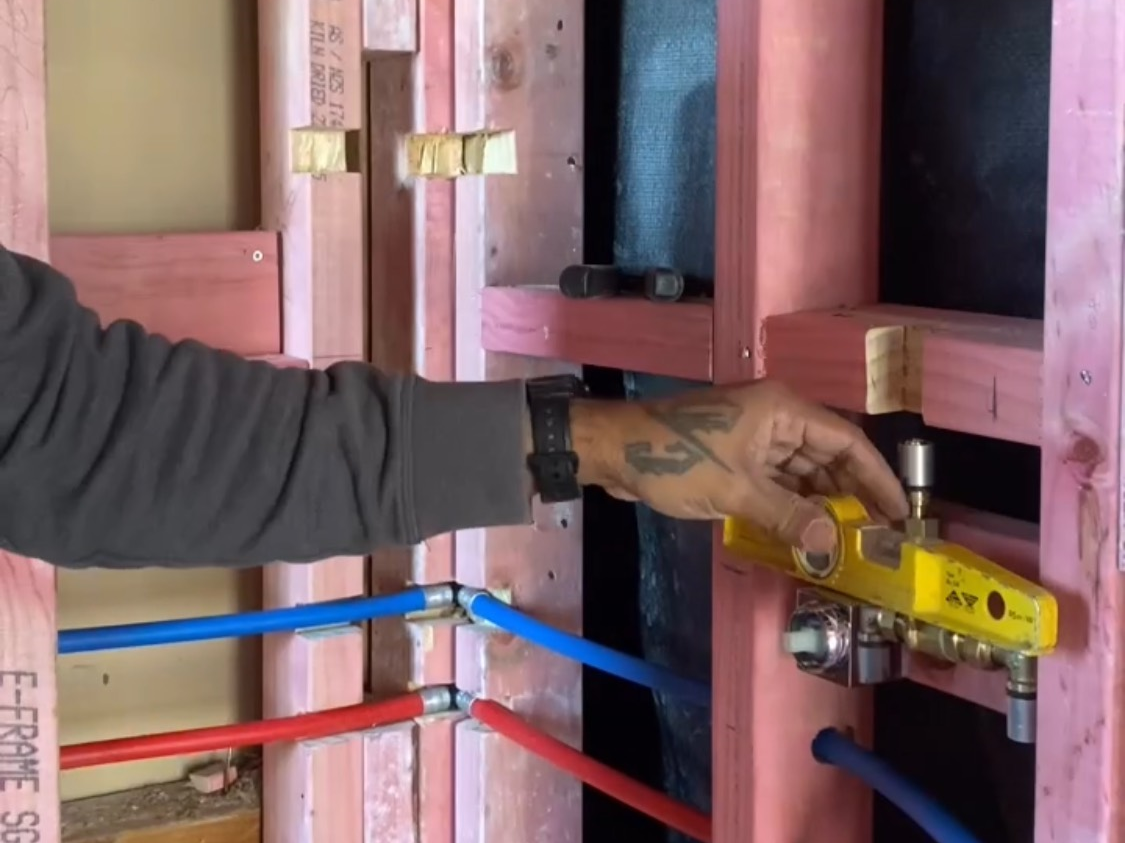 A person using a yellow pneumatic nail gun to fasten a piece of wood in a construction framing project. The person's arm is visible with tattoos and a black watch. The framing is made of pink-colored wood, with plumbing pipes and a level nearby.