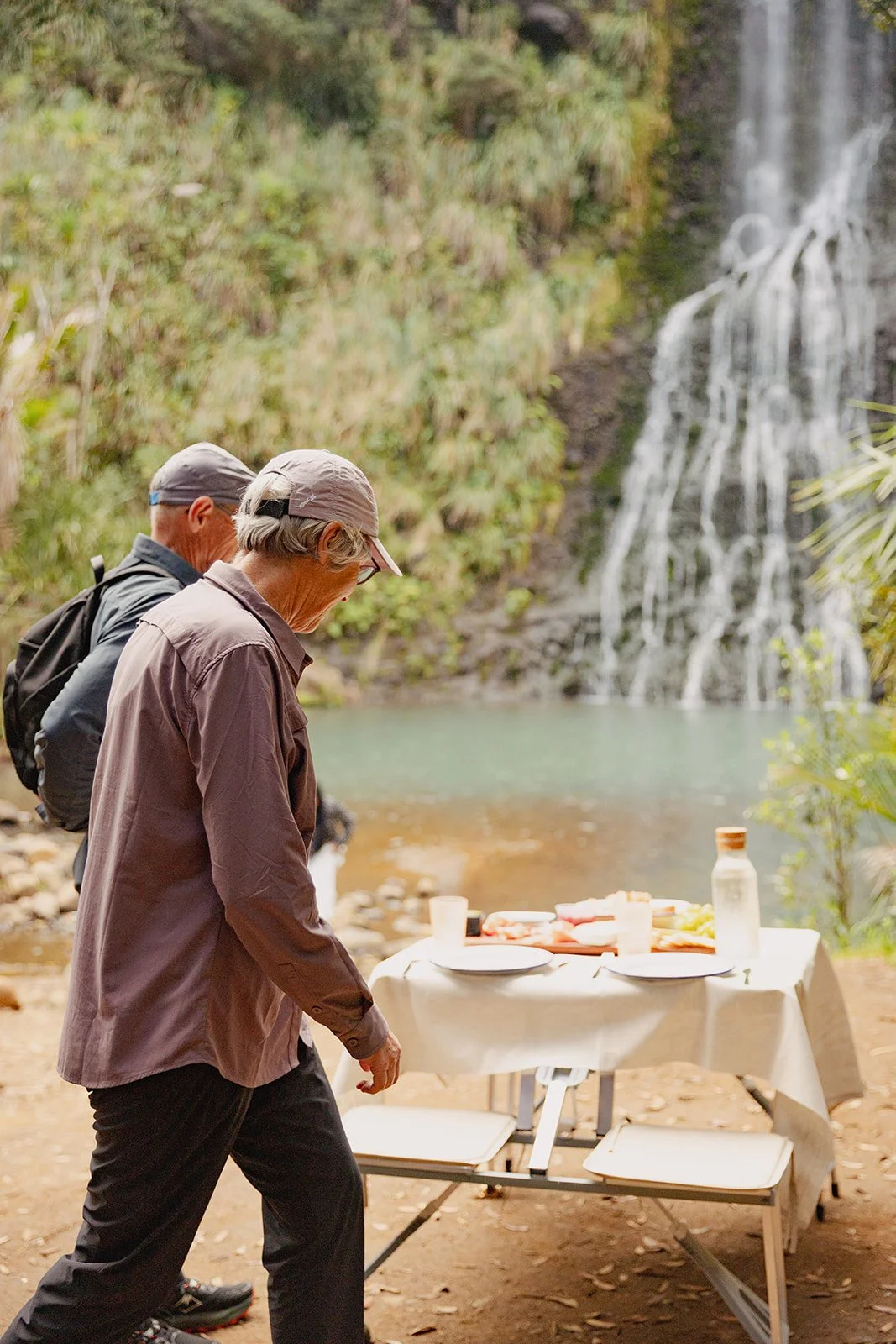 Two older hikers with backpacks standing outdoors near a waterfall and a river, with a picnic table set with food and drinks