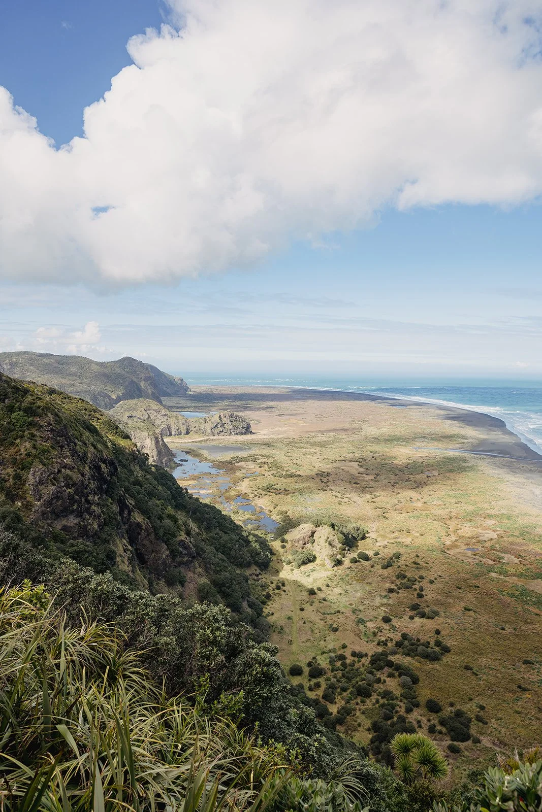 A coastal landscape with green hills in the foreground, a vast flat plain, and the ocean in the distance under cloudy skies.
