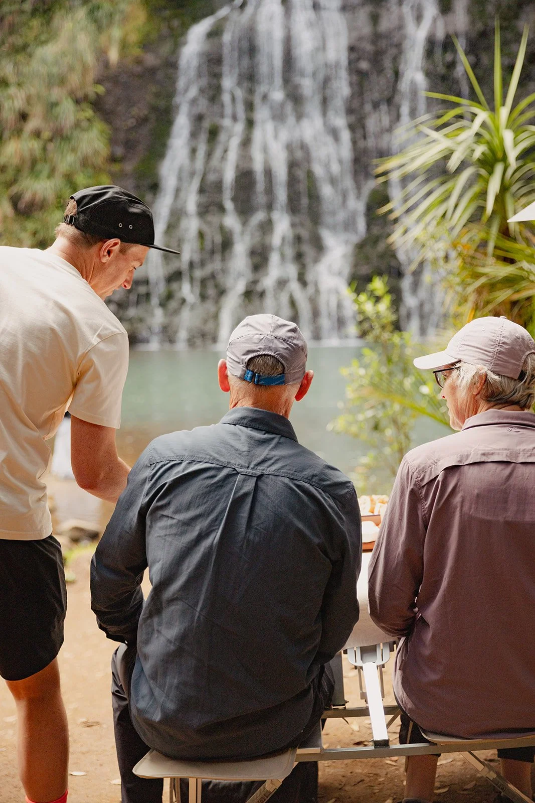 Three elderly people sitting at an outdoor table near a waterfall, with one person standing next to them, in a lush natural setting.
