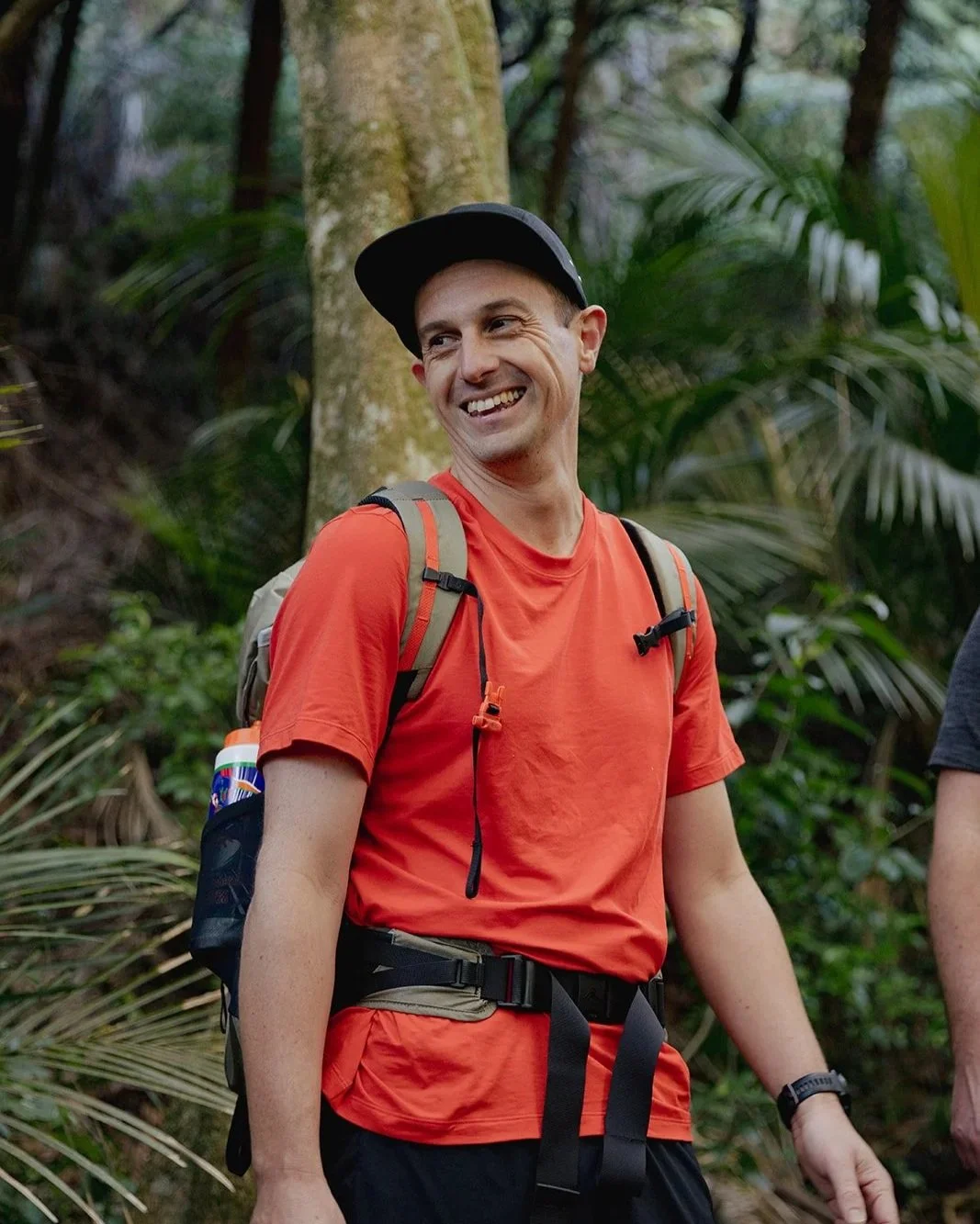A founder guide smiling outdoors in a forest, wearing a red shirt, a black cap, and a backpack.