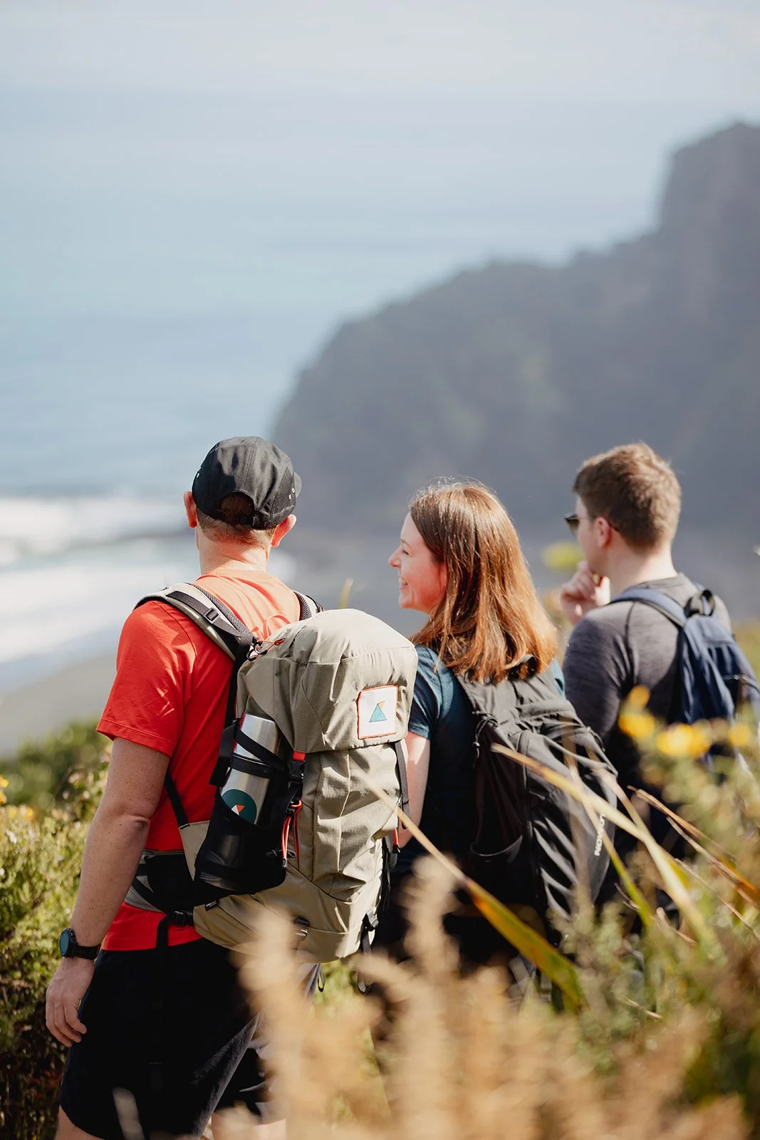 Three hikers standing on a trail overlooking the ocean, with cliffs in the background, all wearing backpacks.