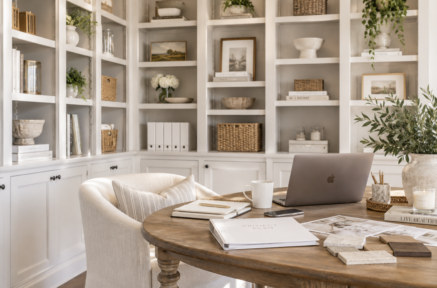 Home office with a round wooden table, a laptop, notebooks, and a white coffee mug, surrounded by white built-in bookshelves filled with books, decorative baskets, paintings, and potted plants, in a cozy and stylish workspace.