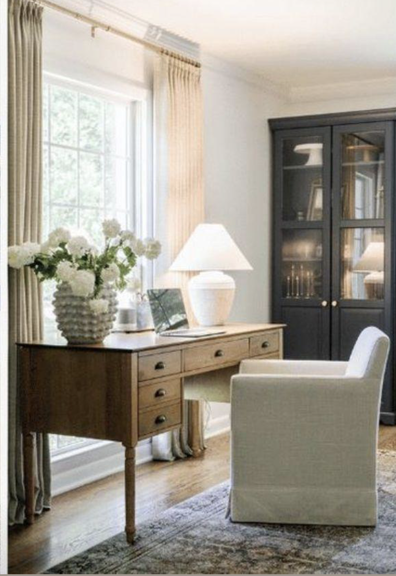 Sunlit cozy corner with a wooden desk and a white upholstered chair in a room with a large window, beige curtains, a white ceramic table lamp, a vase with white flowers, and a black cabinet with glass doors.