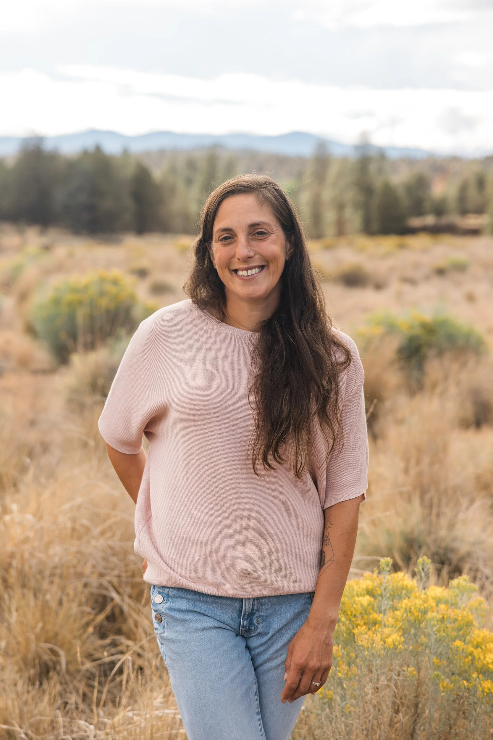 A woman with long dark hair smiling, standing in a field with dry grass and yellow wildflowers, with trees and mountains in the background under a cloudy sky.