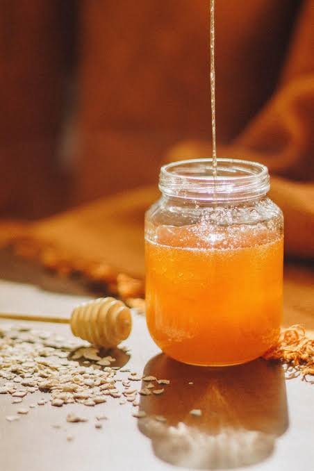 Jar of raw unfiltered honey with honey dipper and honey streak falling into it, surrounded by scattered oats on a wooden surface.