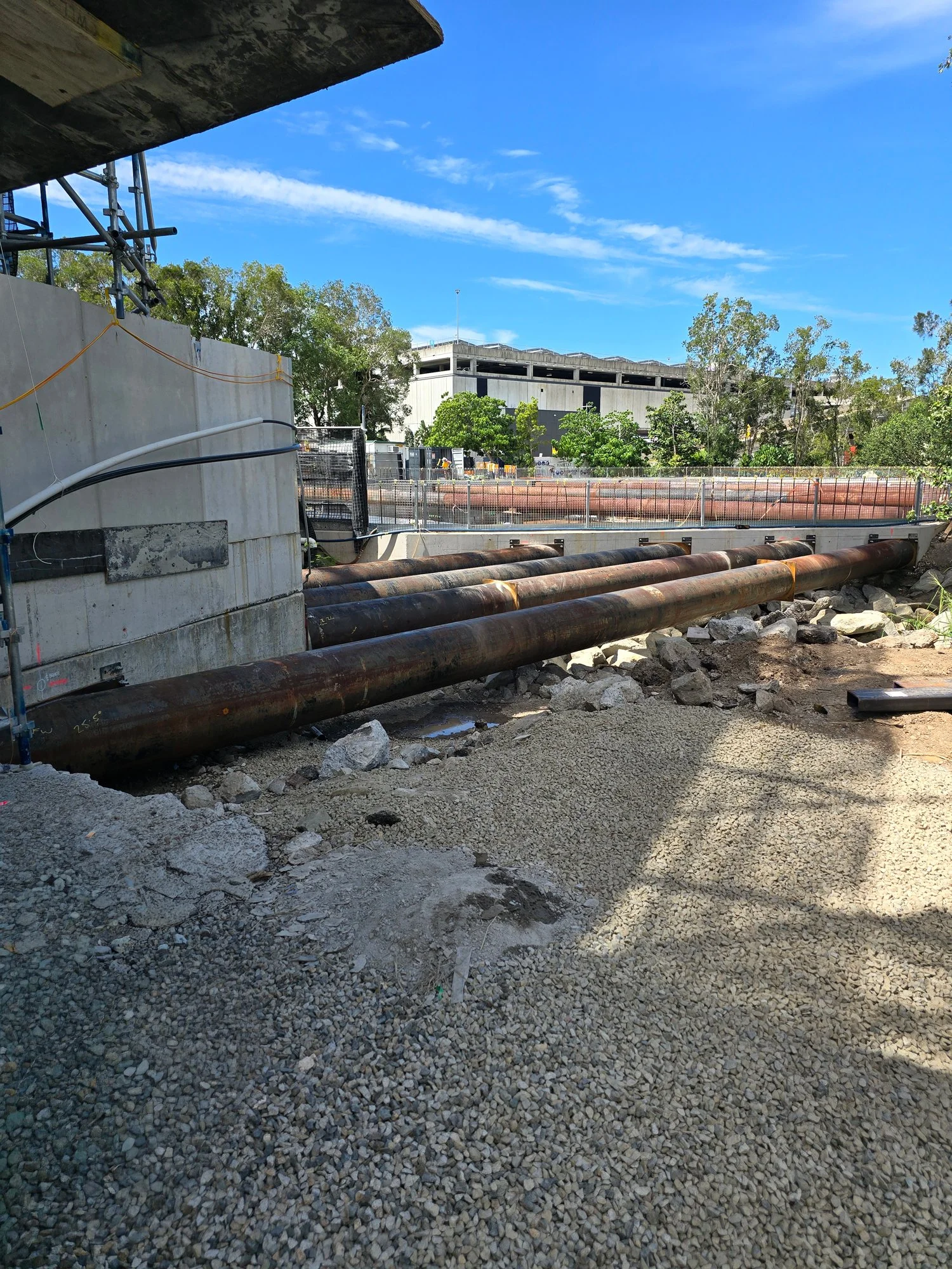 Construction site with large pipes in the foreground, gravel on the ground, a partially built structure, and trees under a blue sky with some clouds.
