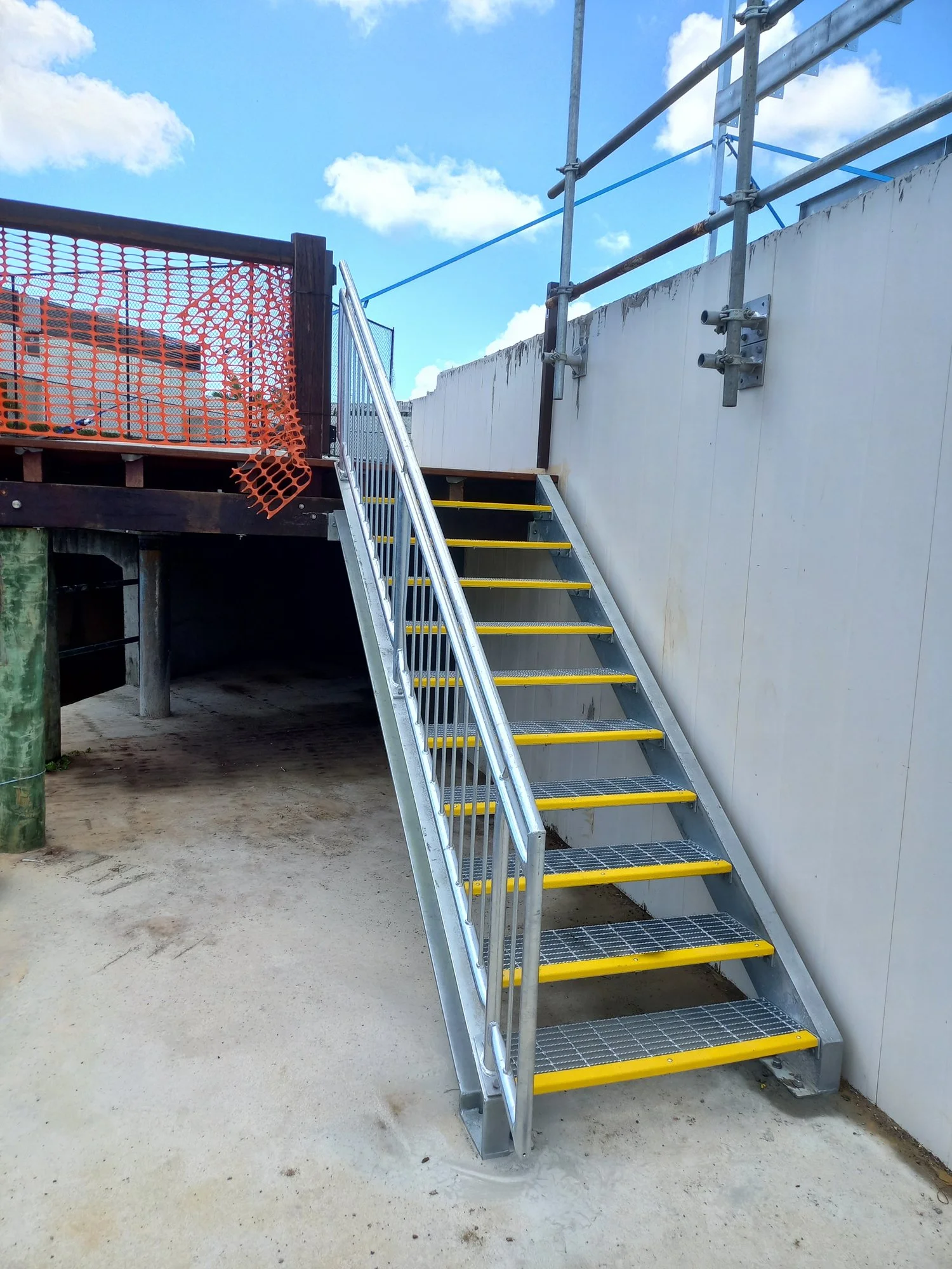 Metal staircase with yellow safety edges leading up to a white wall with scaffolding and orange safety netting nearby, under partly cloudy blue sky.