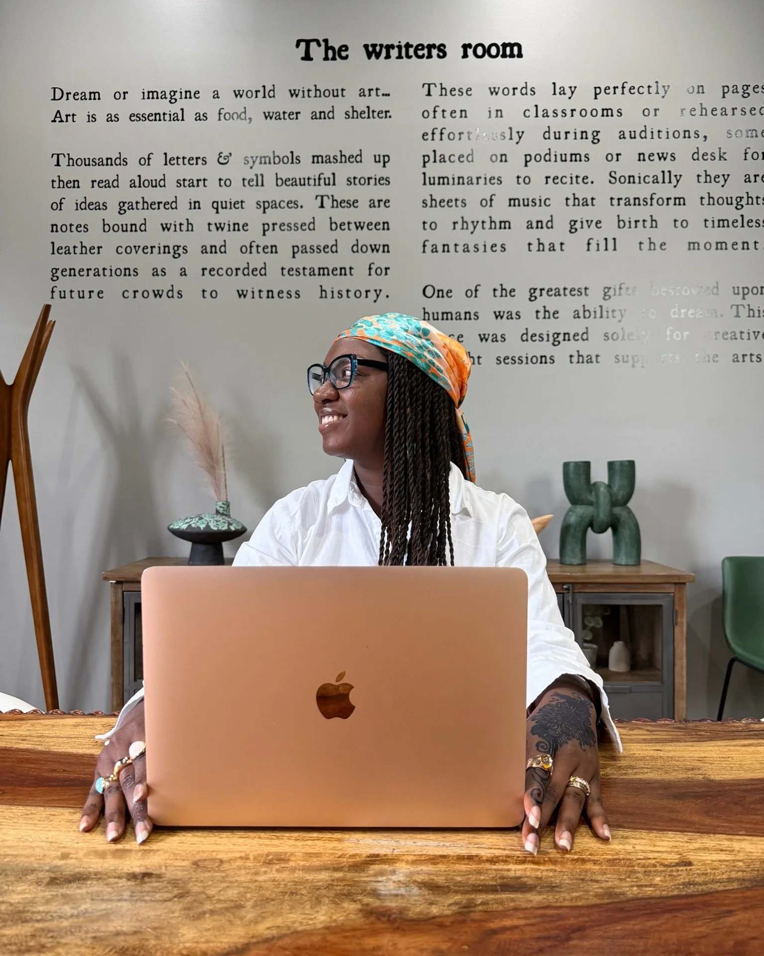 A woman with braids, glasses, and a colorful headscarf sits at a wooden table with a closed rose gold MacBook in front of her, smiling and looking to the side. There is a large white wall with black text behind her titled 'The writers room'.