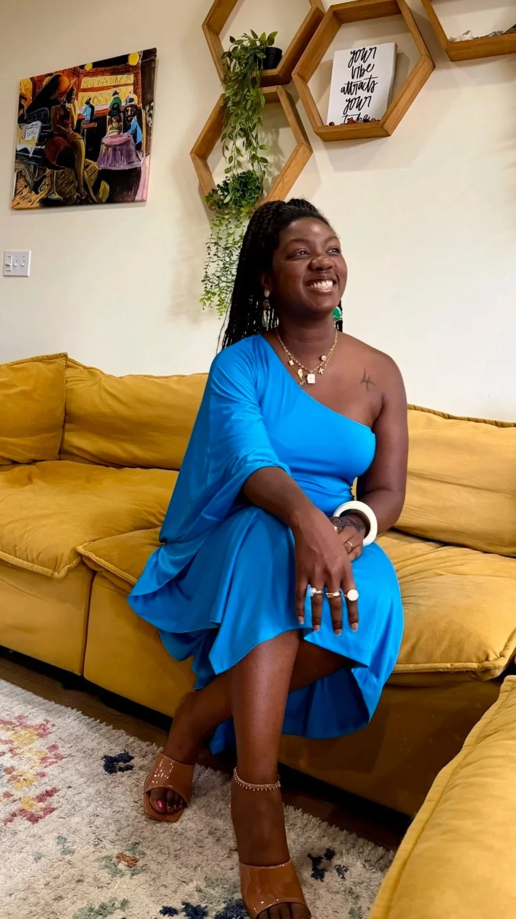 A smiling woman in a blue dress, sitting on a yellow couch, in a living room with wall art and hexagonal wooden shelves with plants.