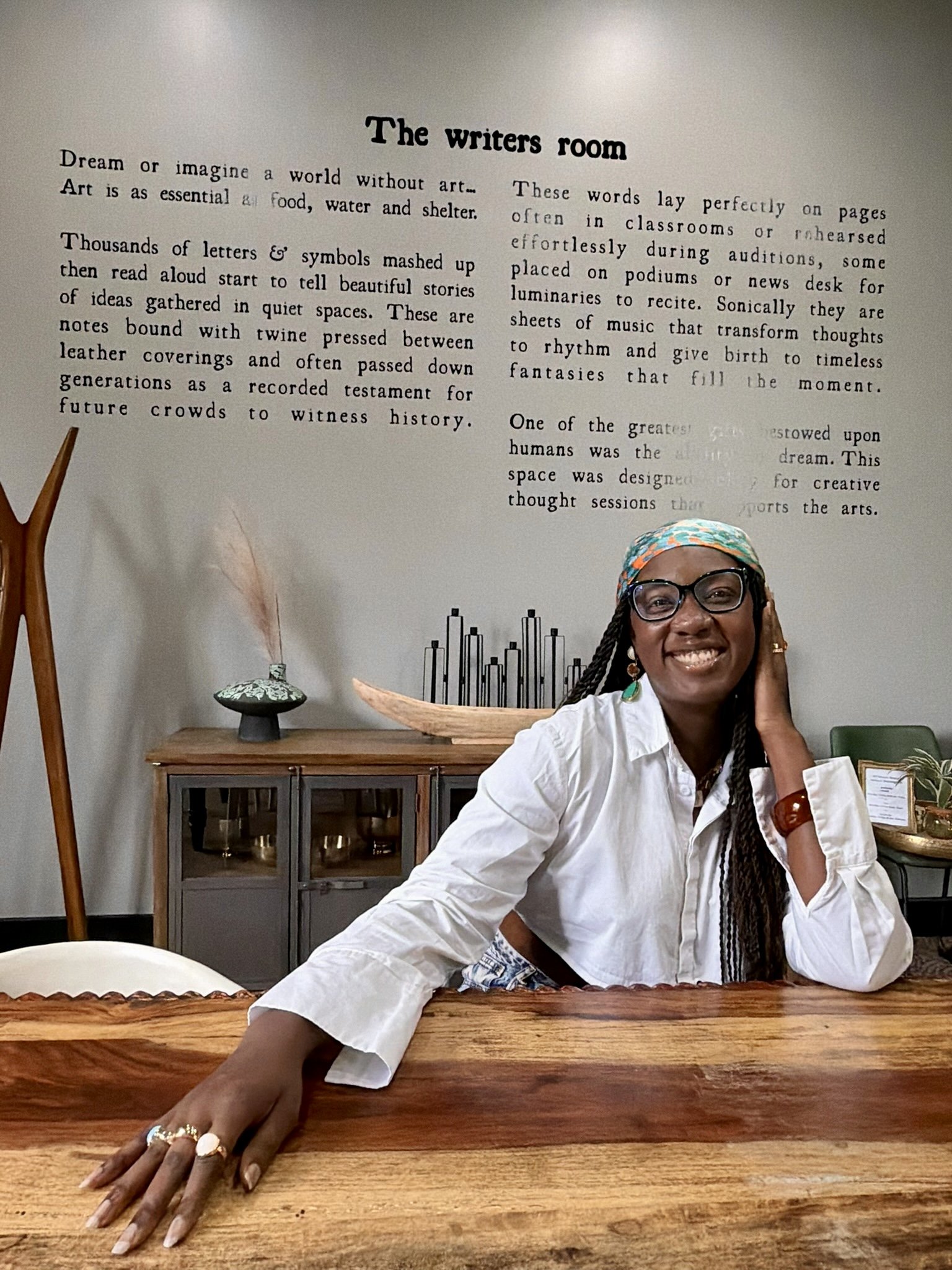 A woman smiling, wearing glasses and a colorful headscarf, sitting at a wooden table in a room with a wall displaying typed text about writers' space and creativity.