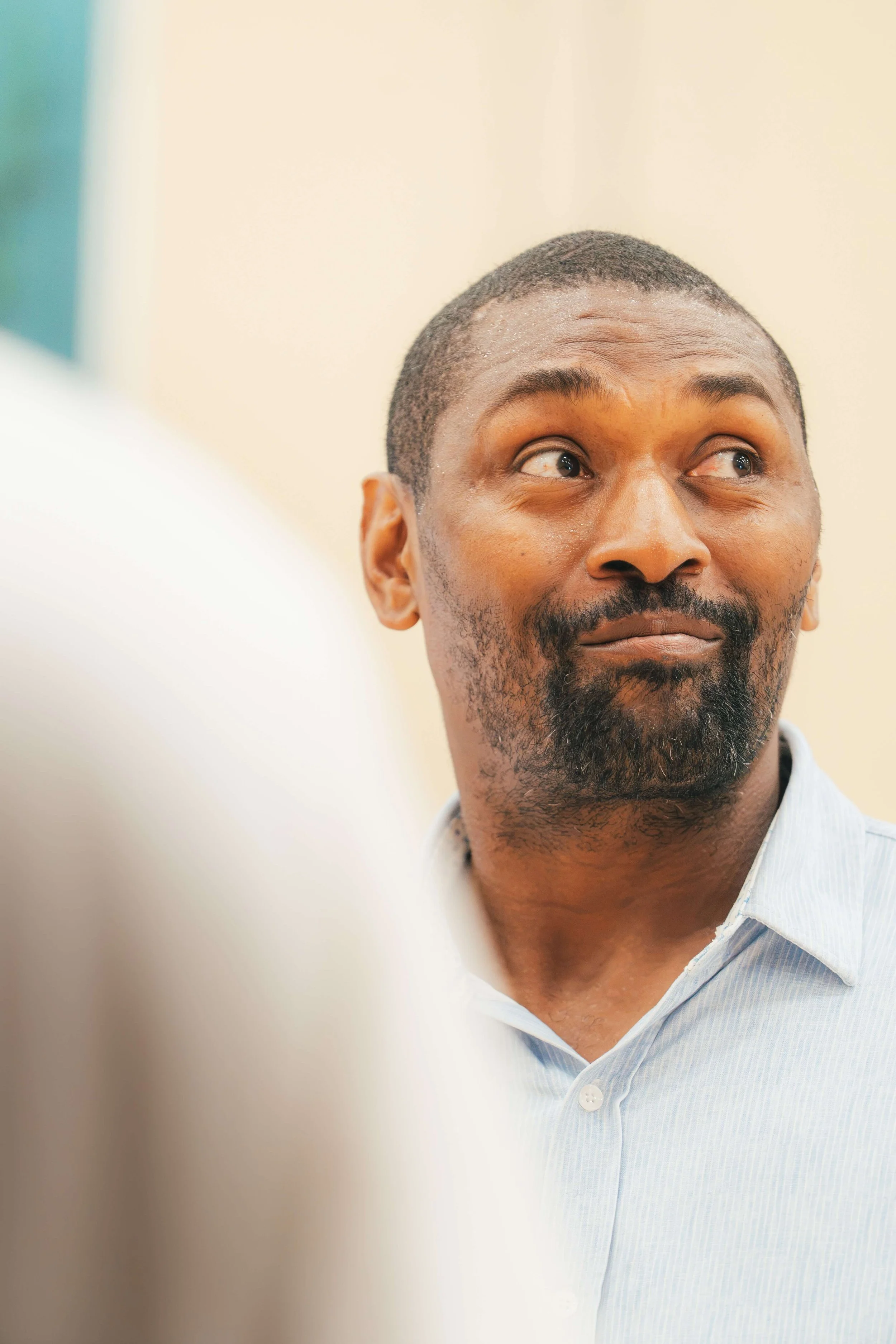 A close-up of Metta World Peace's face with a short haircut and beard, wearing a light blue collared shirt, looking to the side with a slight smile.
