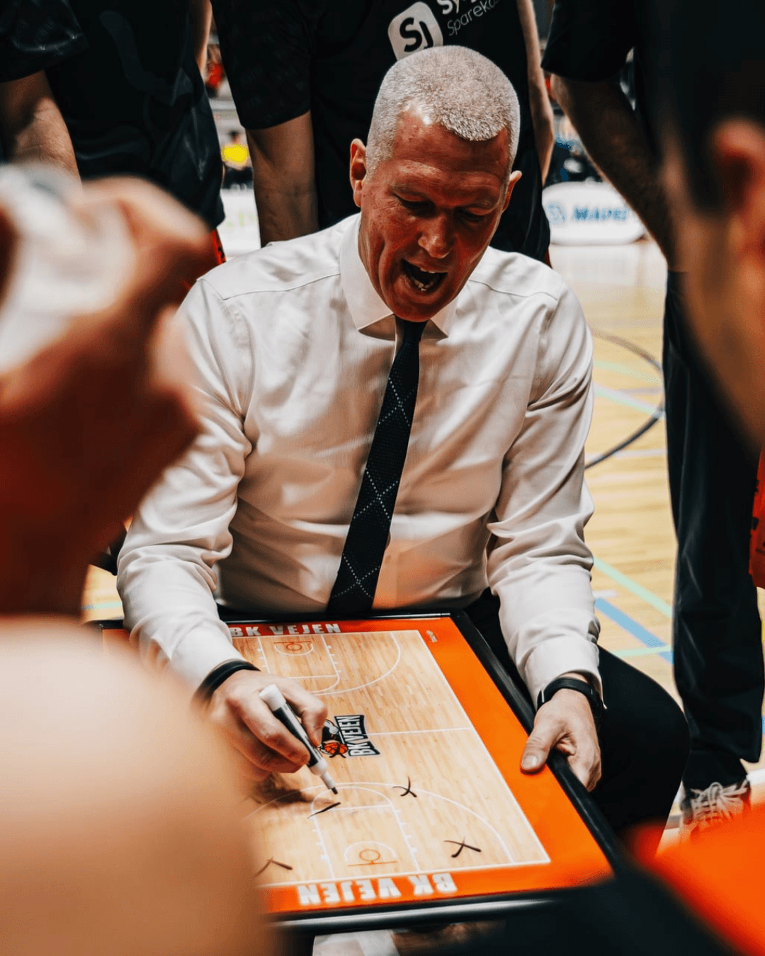 Coach Steve Hanson drawing basketball plays on a clipboard during a timeout, surrounded by his team with intense focus.