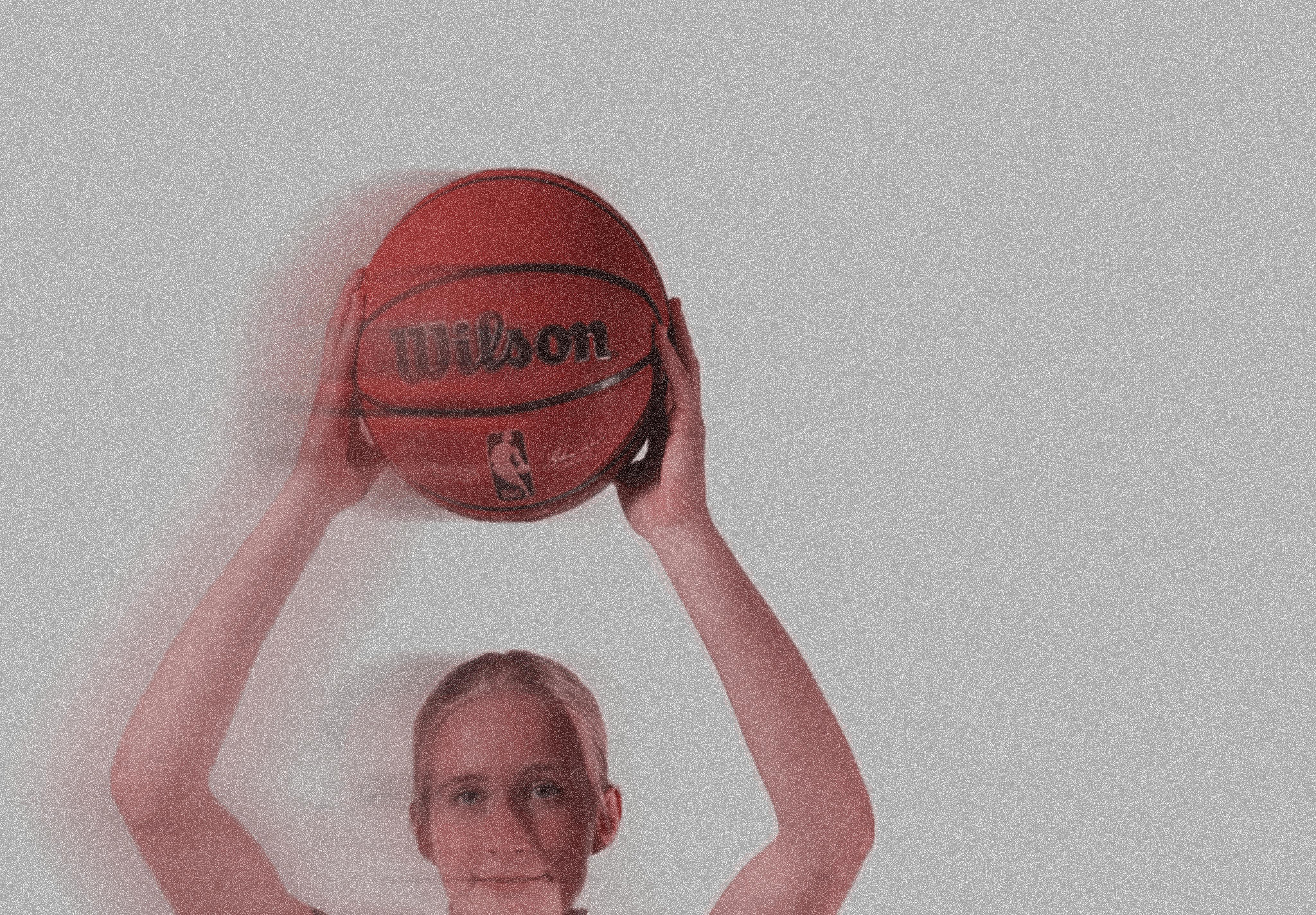 A person holding a red Wilson basketball above their head with both hands, with their face partially visible at the bottom of the image and a dark, textured background.