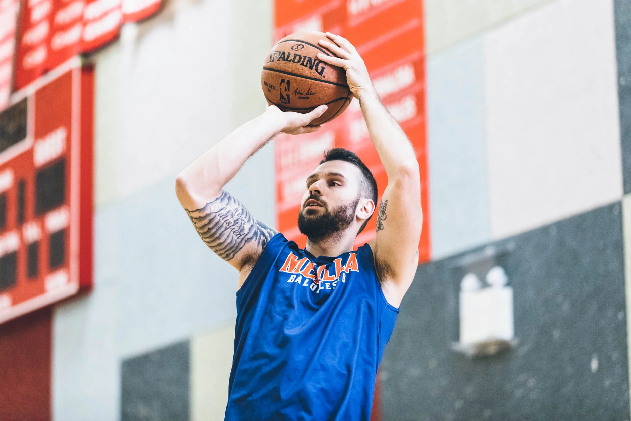 Diego Kapelan in a blue basketball jersey taking a jump shot in an indoor gym, with a red scoreboard on the wall behind him.