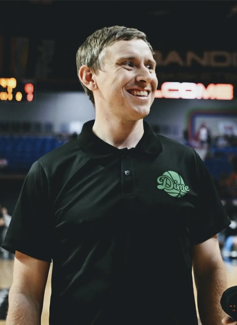 Coach Martin Paul smiling, wearing a black polo shirt with a green logo that says 'Dime' on the chest, standing inside a sports arena or gym.