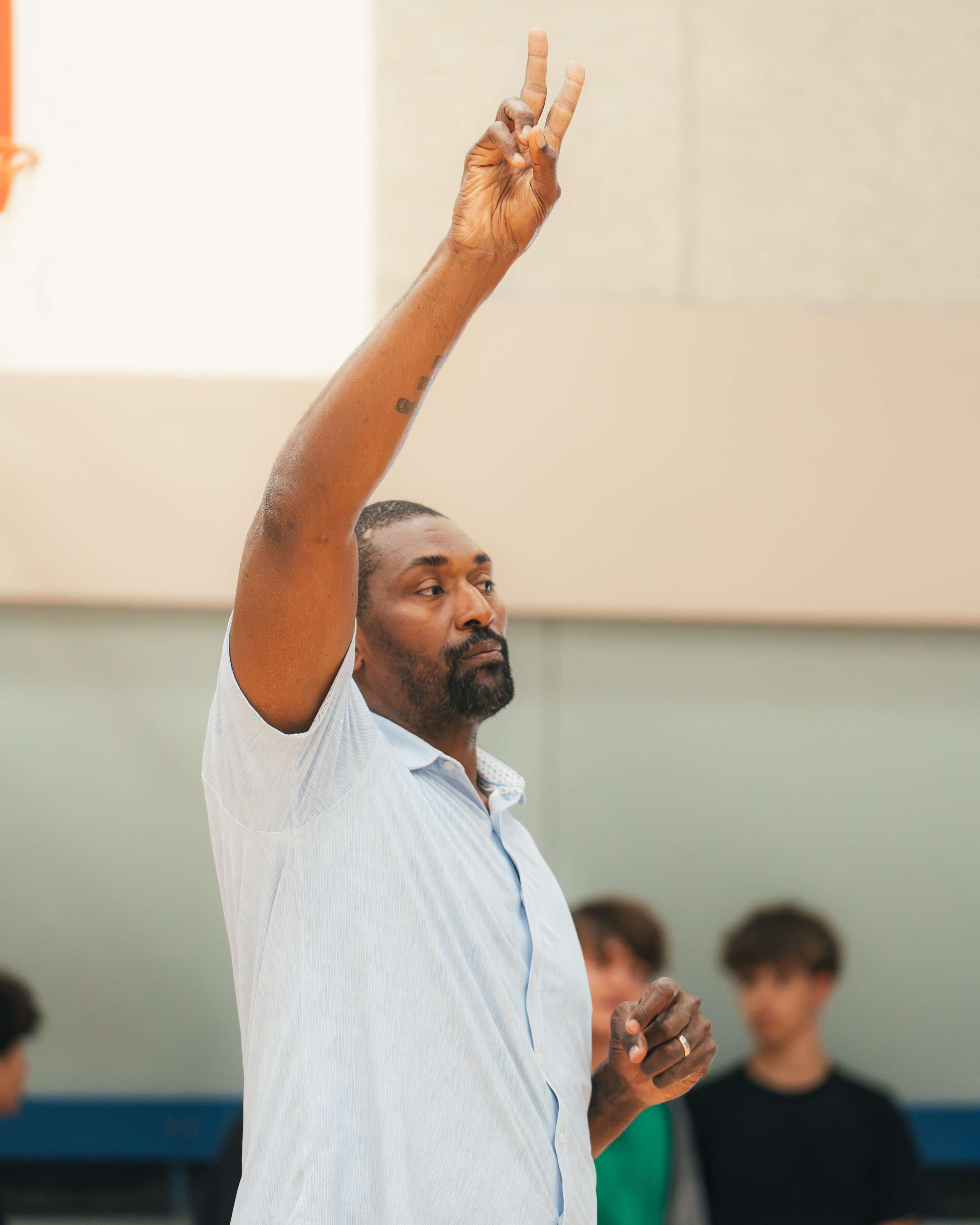 Metta World Peace coaching a gym full of students, holding up two fingers.