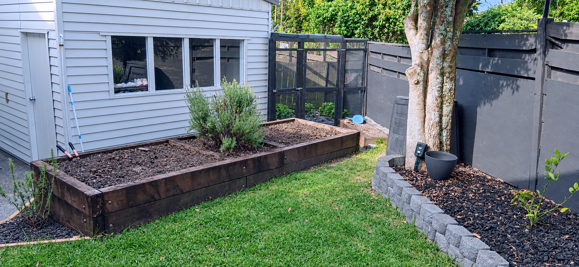 A backyard garden with wooden and concrete raised beds filled with mulch and plants, a small tree, gardening tools, and a black fence with a house in the background.