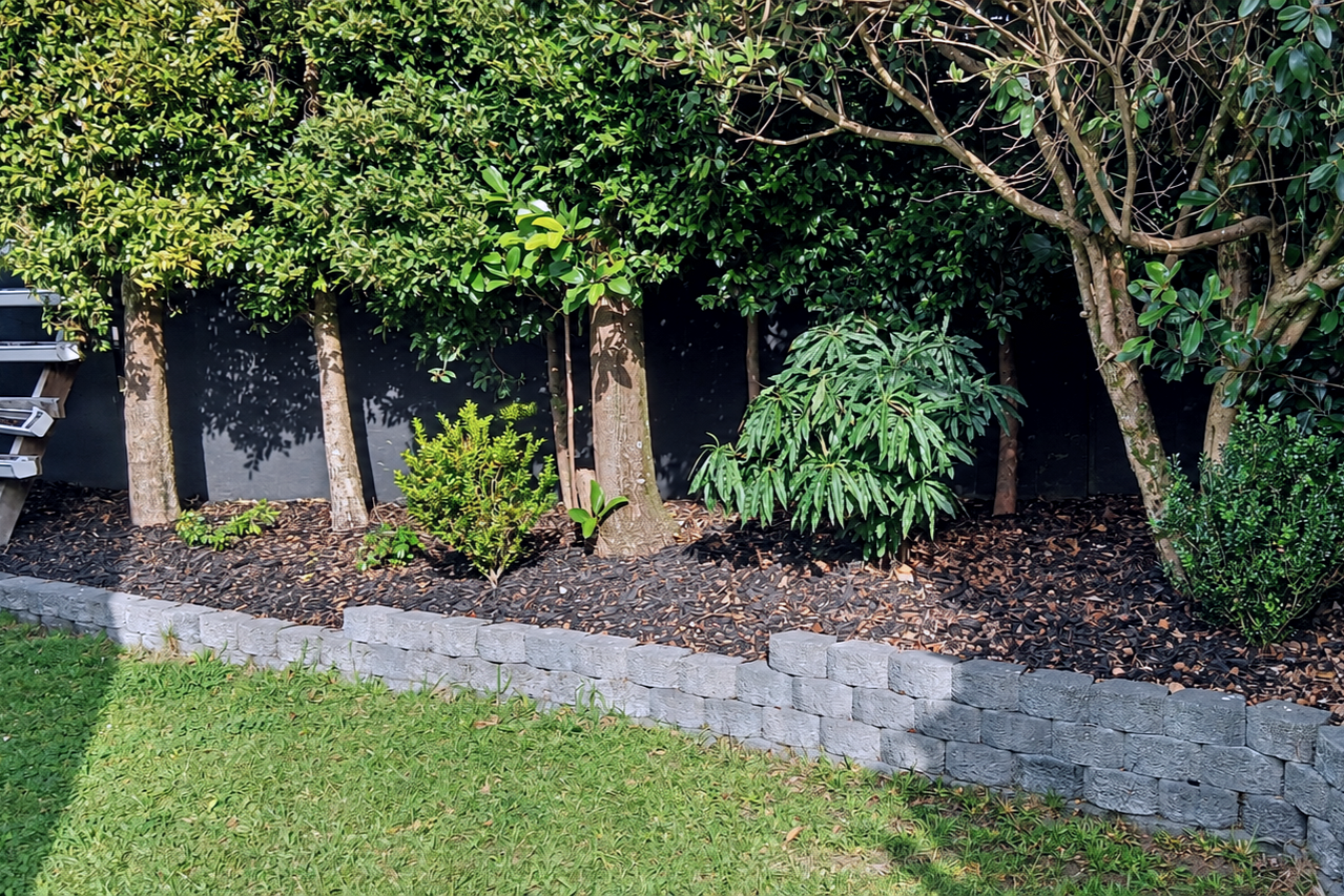 A landscaped garden bed with small trees and shrubs, bordered by gray bricks and covered with mulch, beside a grassy lawn.