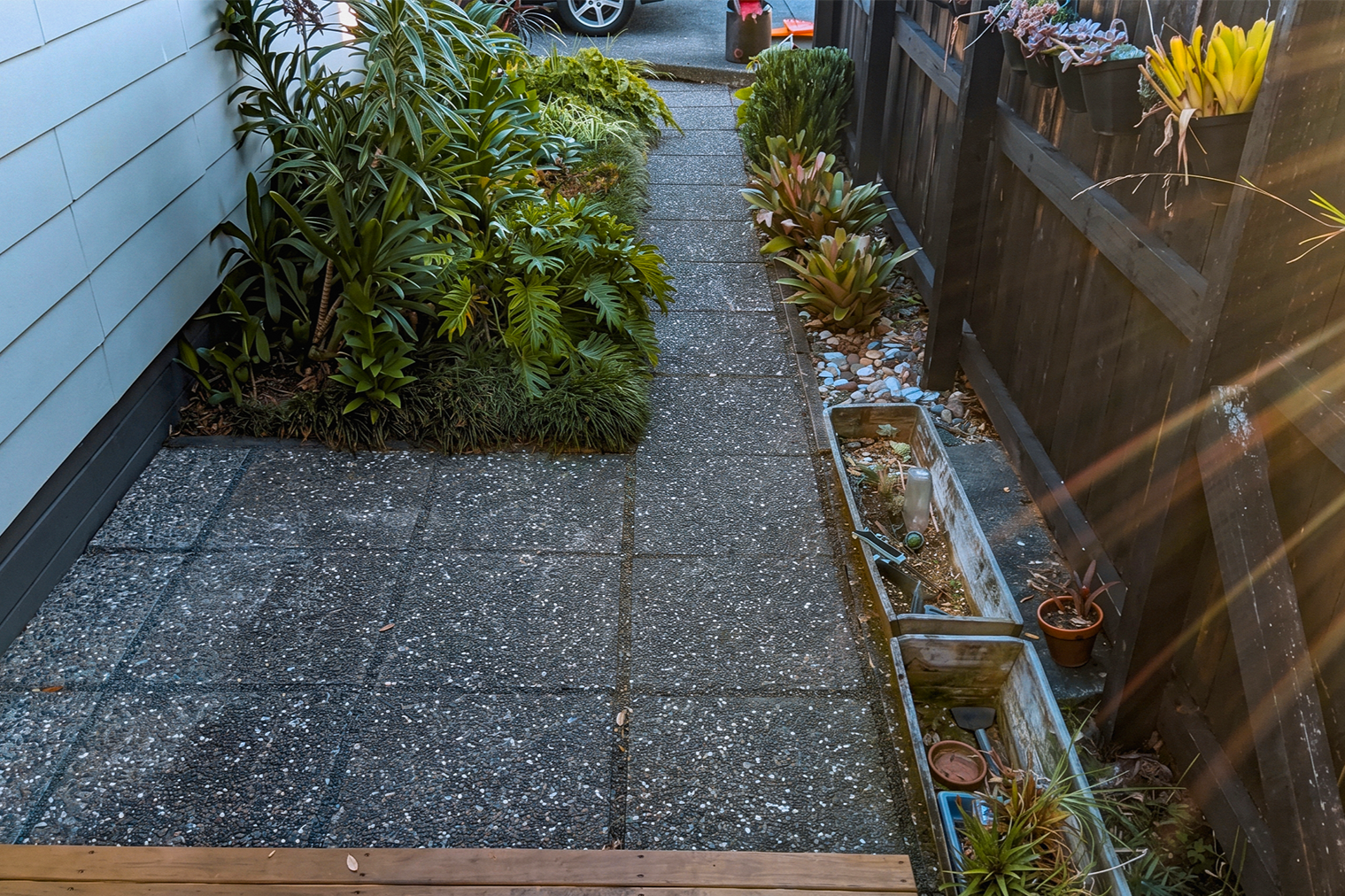 A small backyard patio with textured concrete tiles, bordered by plants on the left and in pots along a black picket fence on the right. Some plant containers and gardening tools are in a narrow planter box on the right.