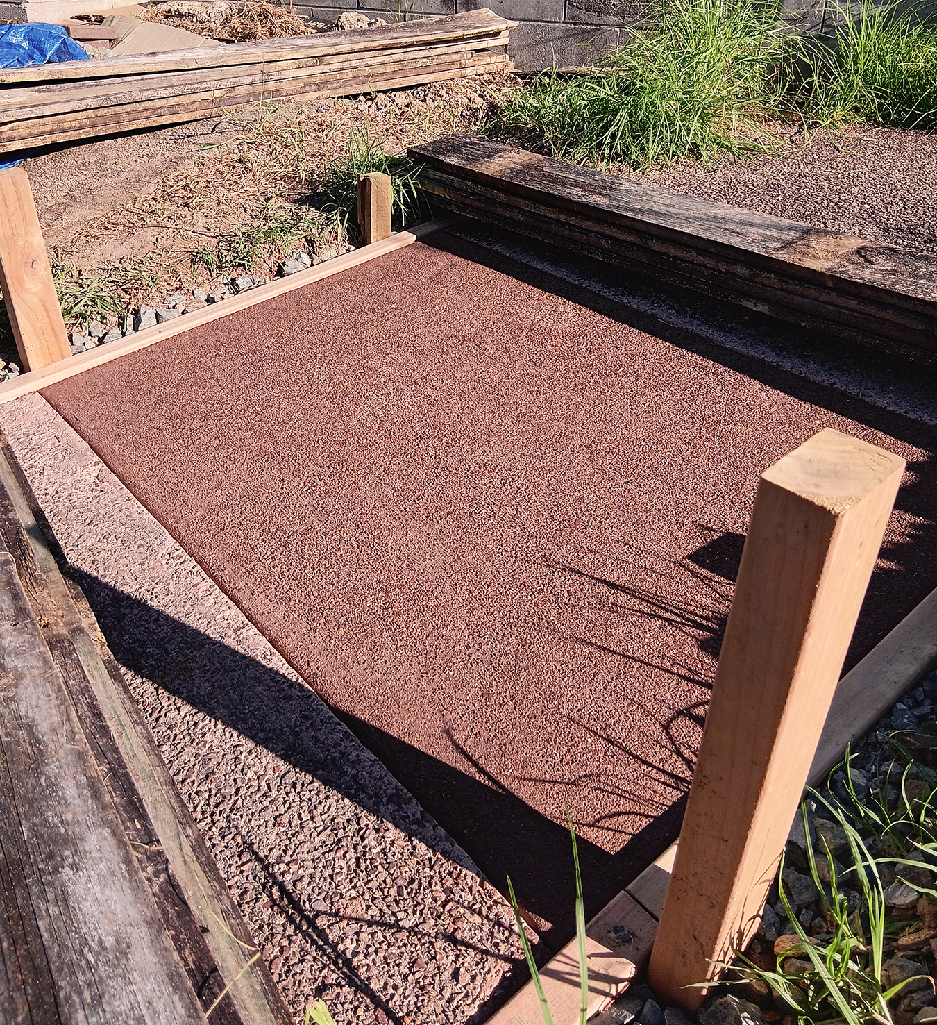A rectangular red textured surface, possibly a hot tub or small pool, under construction, surrounded by wooden planks and greenery.