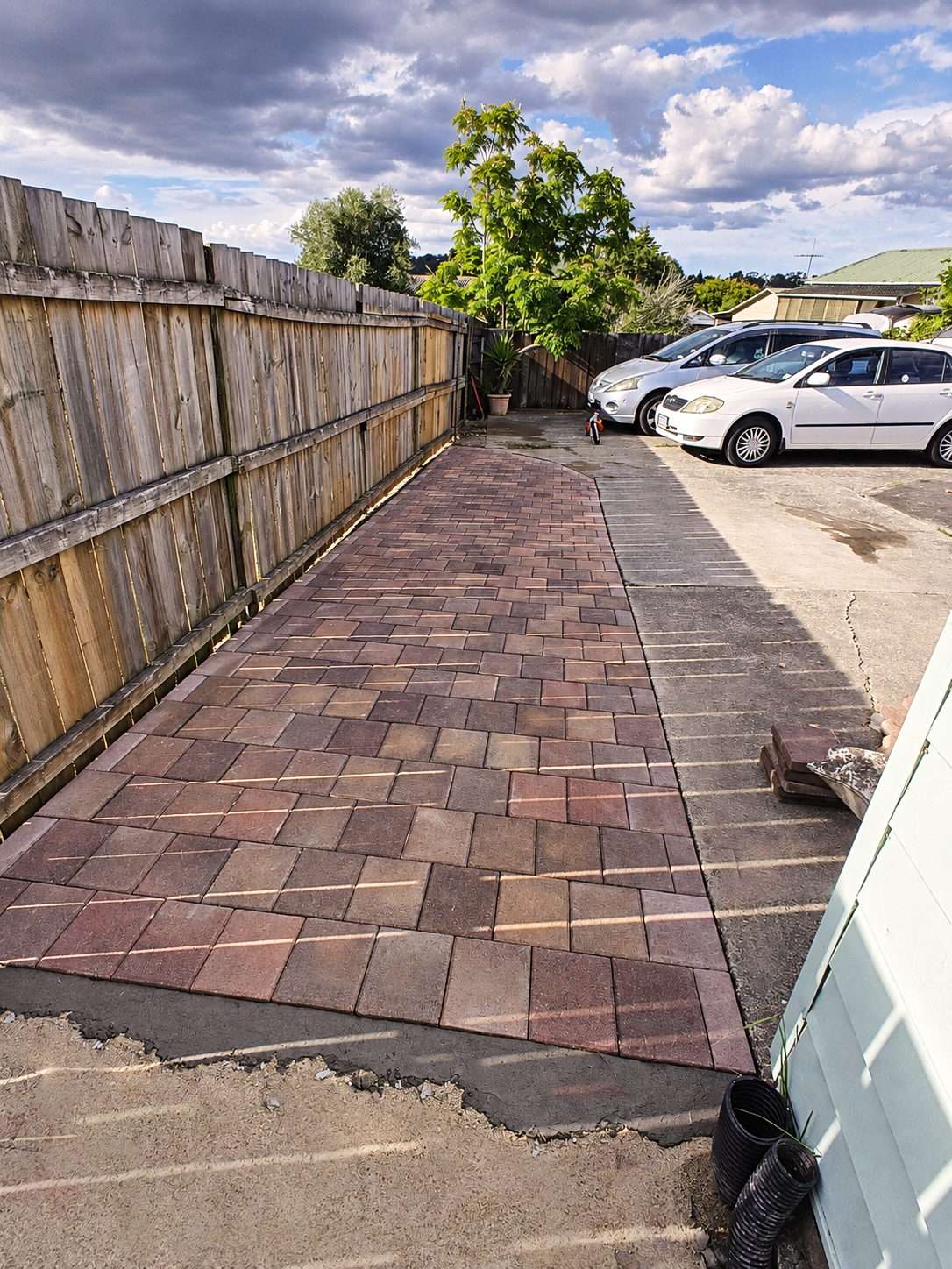 A newly laid brick sidewalk in a residential driveway, with a wooden fence on the left, parked cars on the right, and a large green tree in the background.