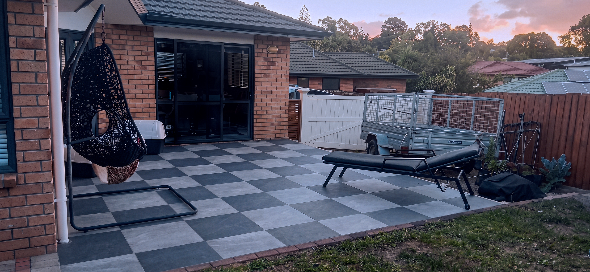 Backyard with checkered tile patio, hanging egg chair, lounge chair, small trailer, fence, and sliding glass door at sunset.