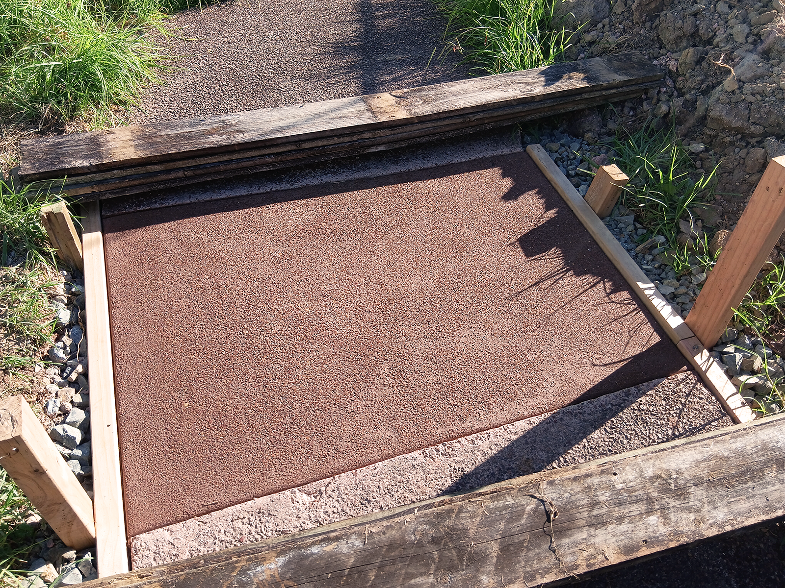A miniature train crossing with a textured, reddish-brown track surface, framed by wooden rails, with grass and gravel on the sides.