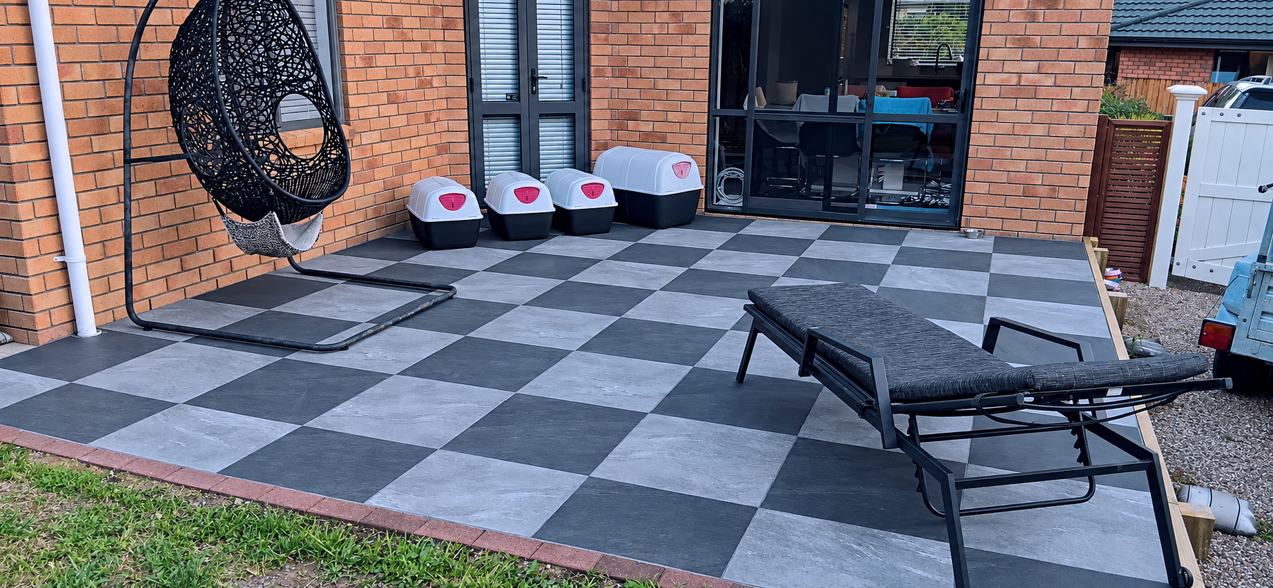 A backyard patio with black and white checkered tiles, a black outdoor lounge chair, a black hanging egg chair, four pet carriers with pink handles, and a glass sliding door leading into a house. There is a wooden fence and some outdoor furniture vis