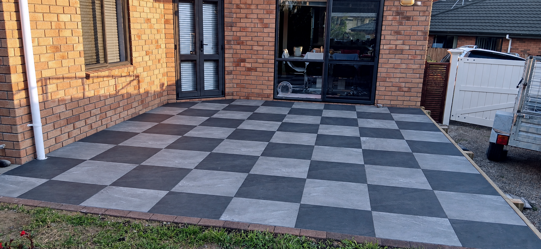 A newly paved outdoor deck with a black and gray checkered tile pattern, attached to a brick house with large glass doors and windows, and a white gate on the right side, with some construction materials still present.