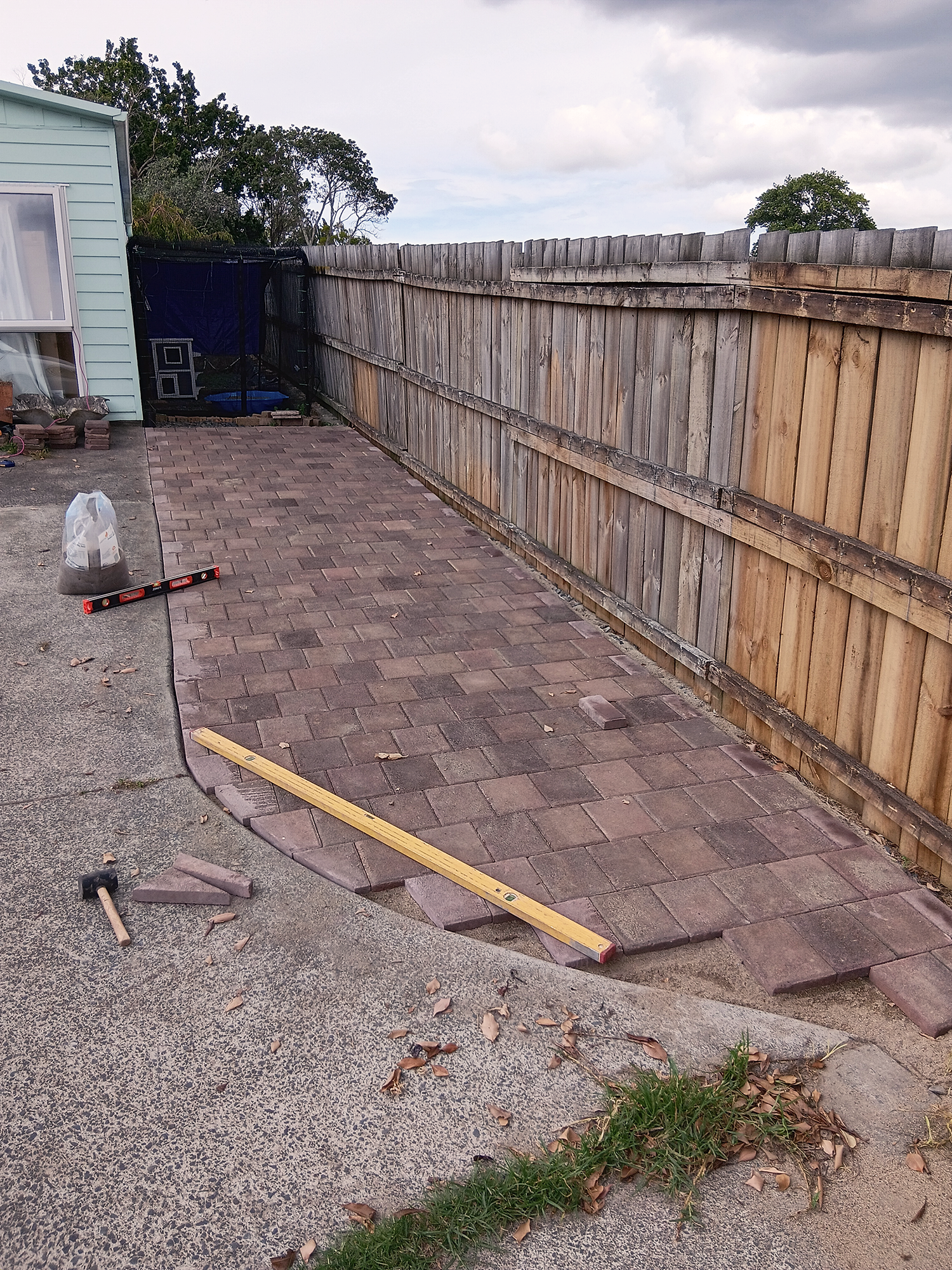 A backyard with a partially completed brick patio, a wooden fence, a small patch of grass, a house with light blue siding, and outdoor tools and materials. There is a black mesh enclosure in the backyard, and some clouds in the sky.
