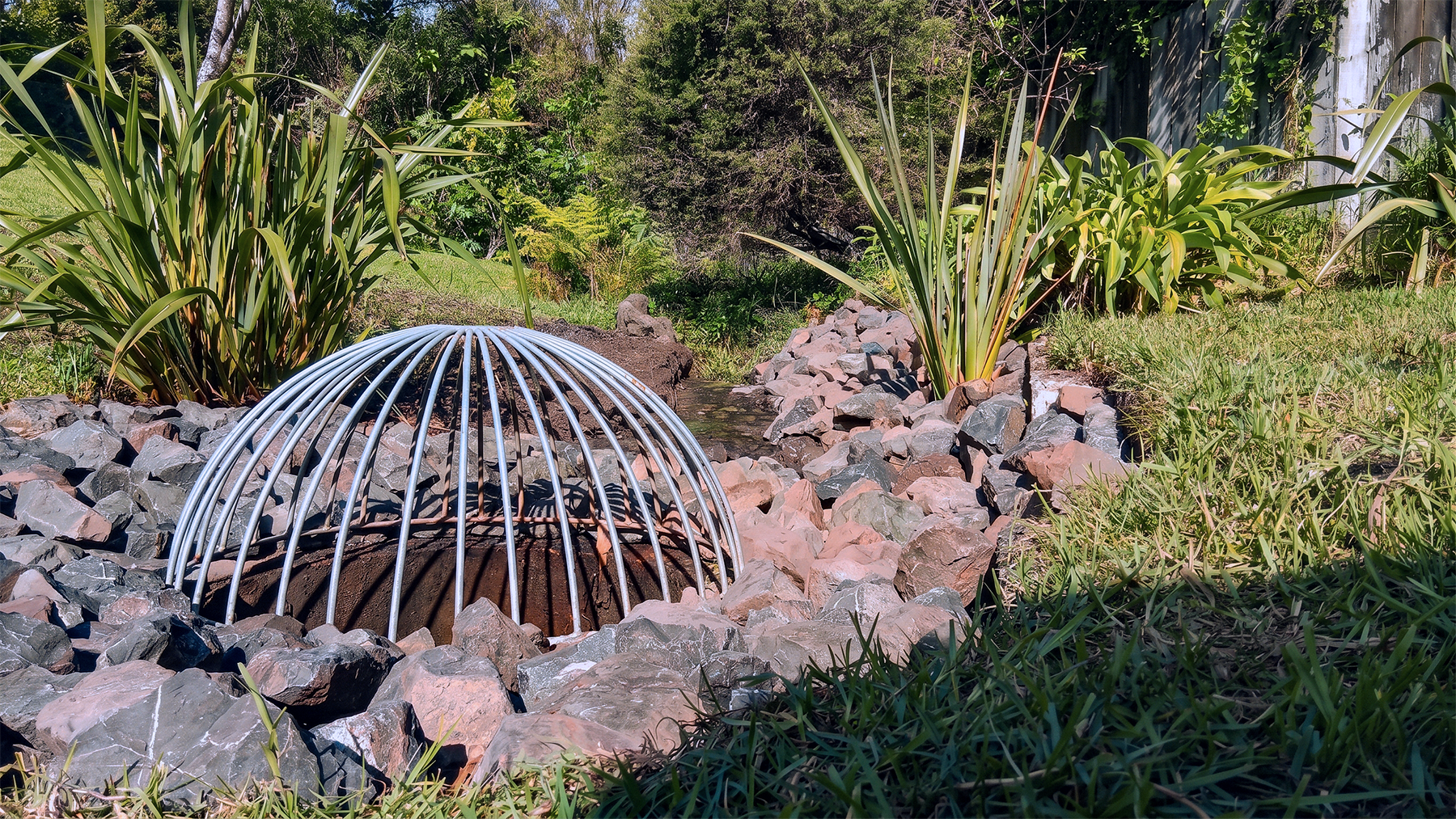 A garden with rocks, green plants, and bushes surrounding a metal dome structure on the ground near a small stream.