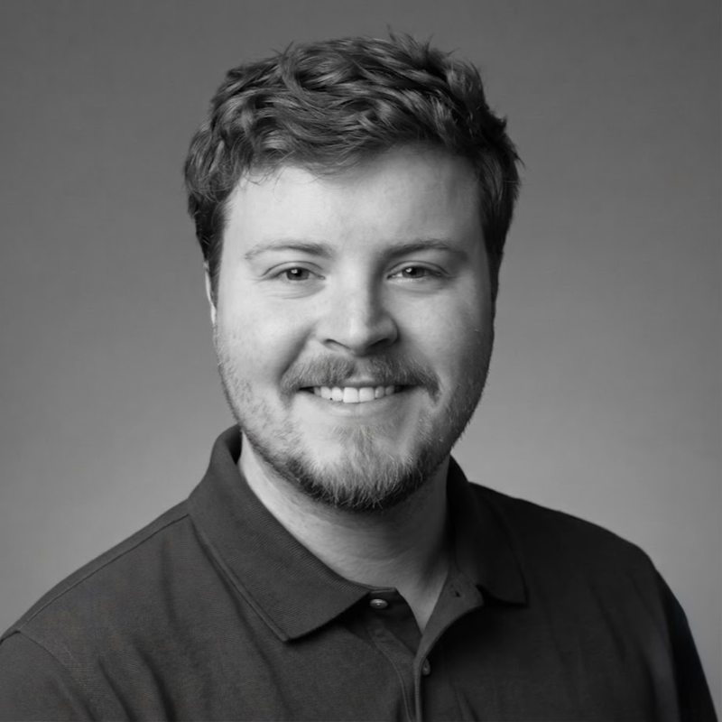 Black and white portrait of a smiling young man with short, curly hair and a beard, wearing a collared shirt.
