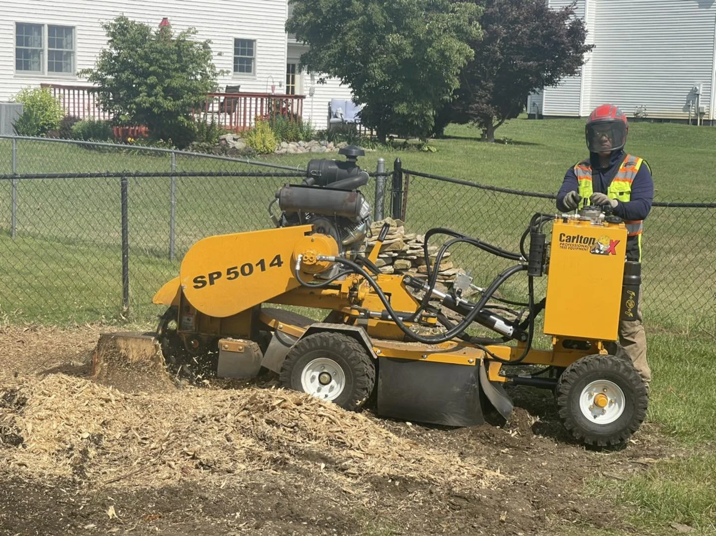 Traditions Tree Care professional crew operating a yellow wood cutting machine outdoors on a grassy area, with a residential house and trees in the background.