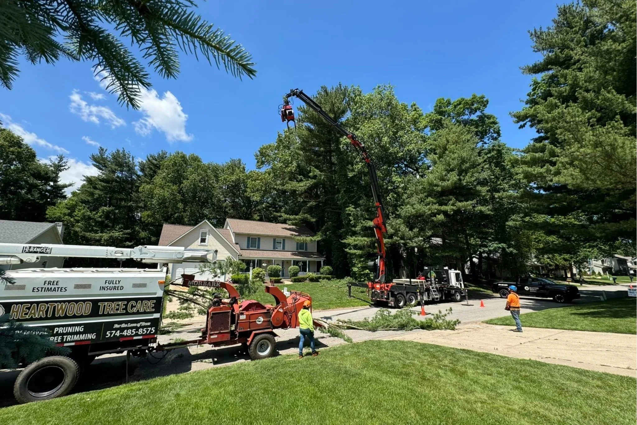 Traditions Tree trimming in a suburban neighborhood with workers and machinery, including a crane lifting a large tree branch, under a clear blue sky.