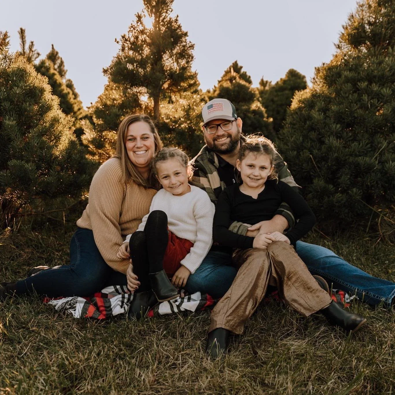 The owners of Traditions and their Family of four sitting on grass outdoors at sunset, surrounded by trees, smiling at the camera.