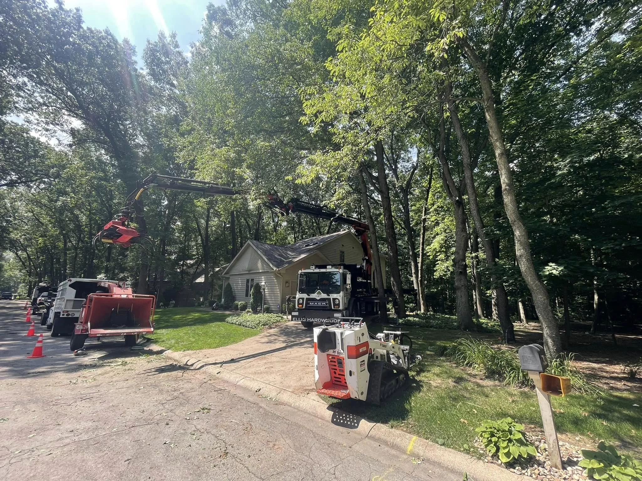 Traditions tree-trimming crew is working in a residential neighborhood, involving a cherry picker lift and a truck with a crane. The scene takes place in a leafy yard with a house, a sidewalk, and parked vehicles, under bright sunlight.