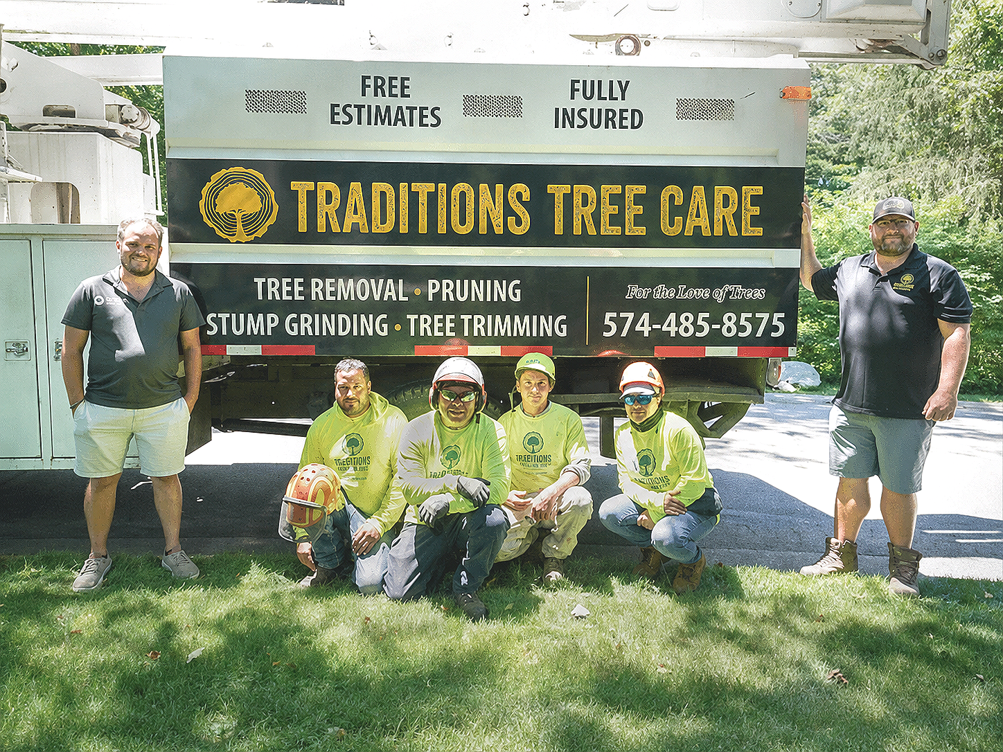 Six people in safety uniforms and two men in business attire posing in front of a Traditions tree care services Truck. The truck has signage for free estimates, fully insured, and a phone number, with the business name 'Traditions Tree Care'.