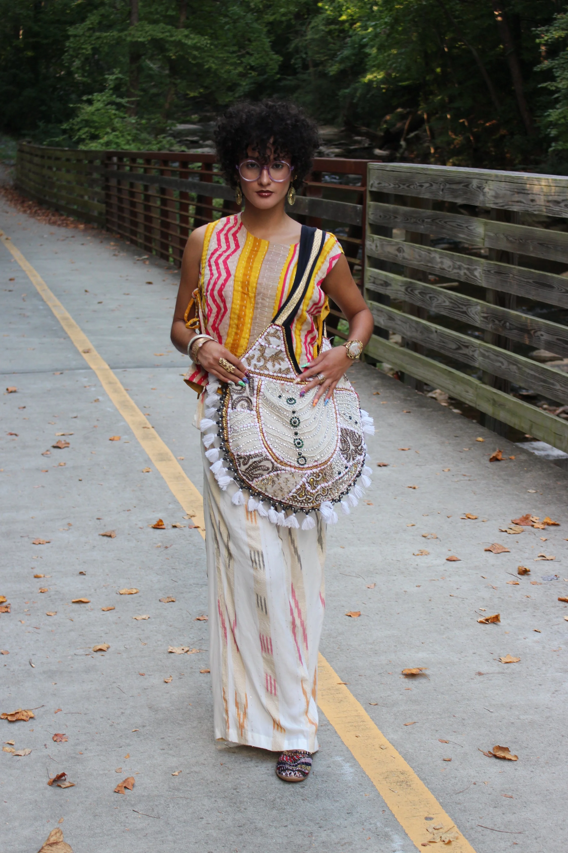 A woman with curly black hair, wearing sunglasses, earrings, and a colorful, patterned sleeveless top, walking on a paved path with fallen leaves, next to a wooden fence and greenery.