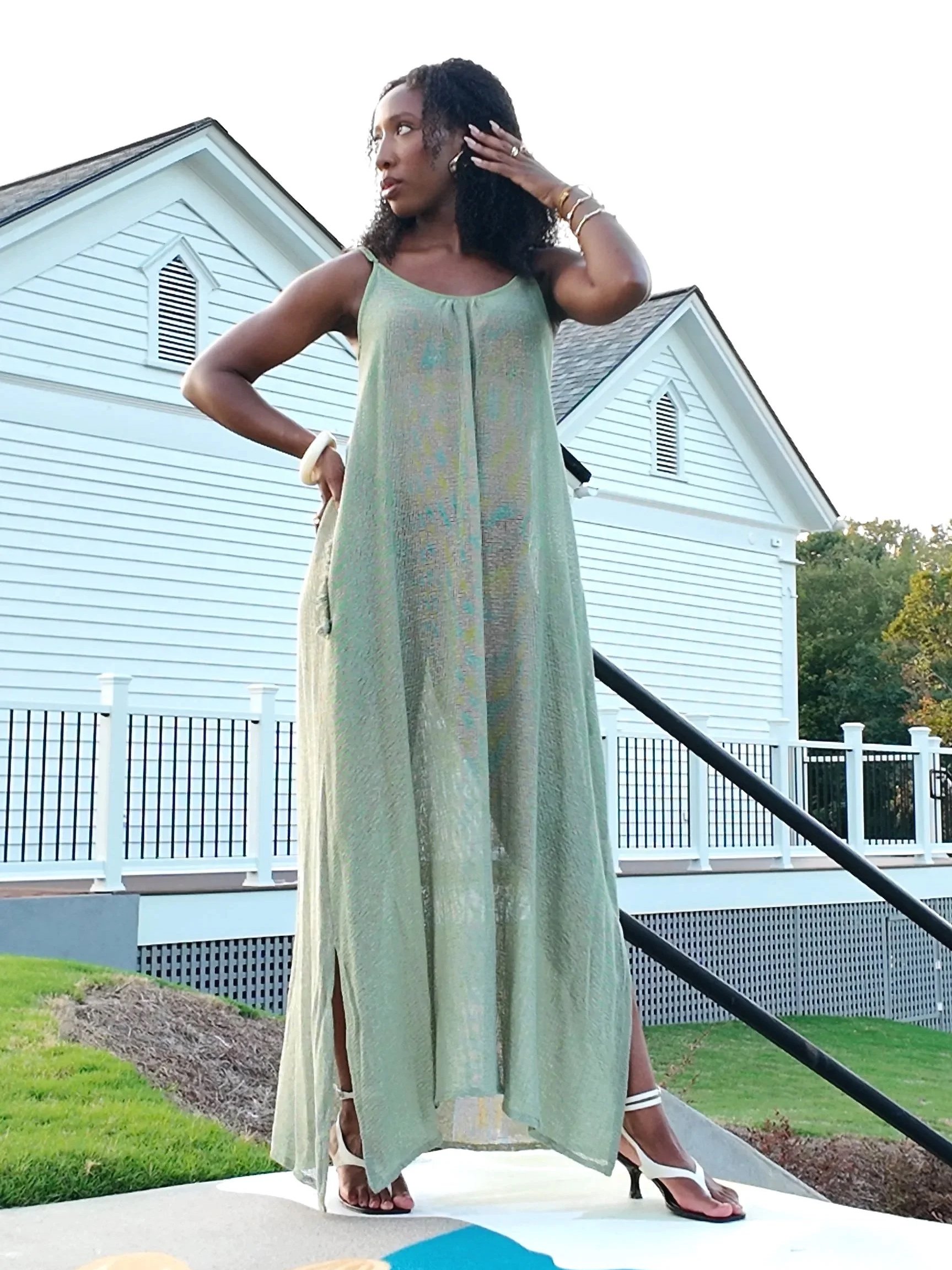A woman with curly black hair, wearing a long, flowing, sheer dress with a pale green hue, stands outdoors on a platform in front of white houses with peaked roofs. She is looking to her left, touching her hair, and is wearing high heels and jewelry.