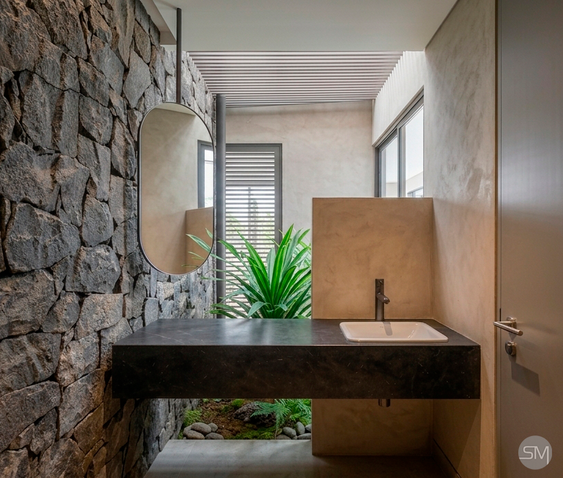 Modern bathroom with stone accent wall, oval mirror, black countertop with white sink, small plant, and windows bringing in natural light.