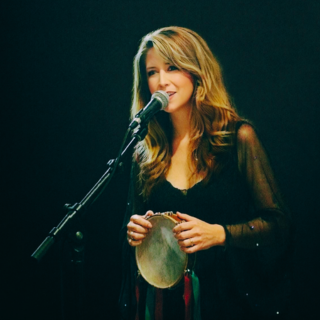 A woman with long wavy hair singing into a microphone while holding a tambourine on a dark stage.