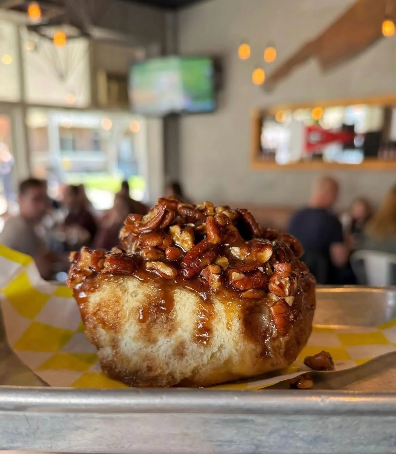 Close-up of a glazed cinnamon roll topped with pecans in a bakery or cafe setting, with blurred customers and a TV in the background.