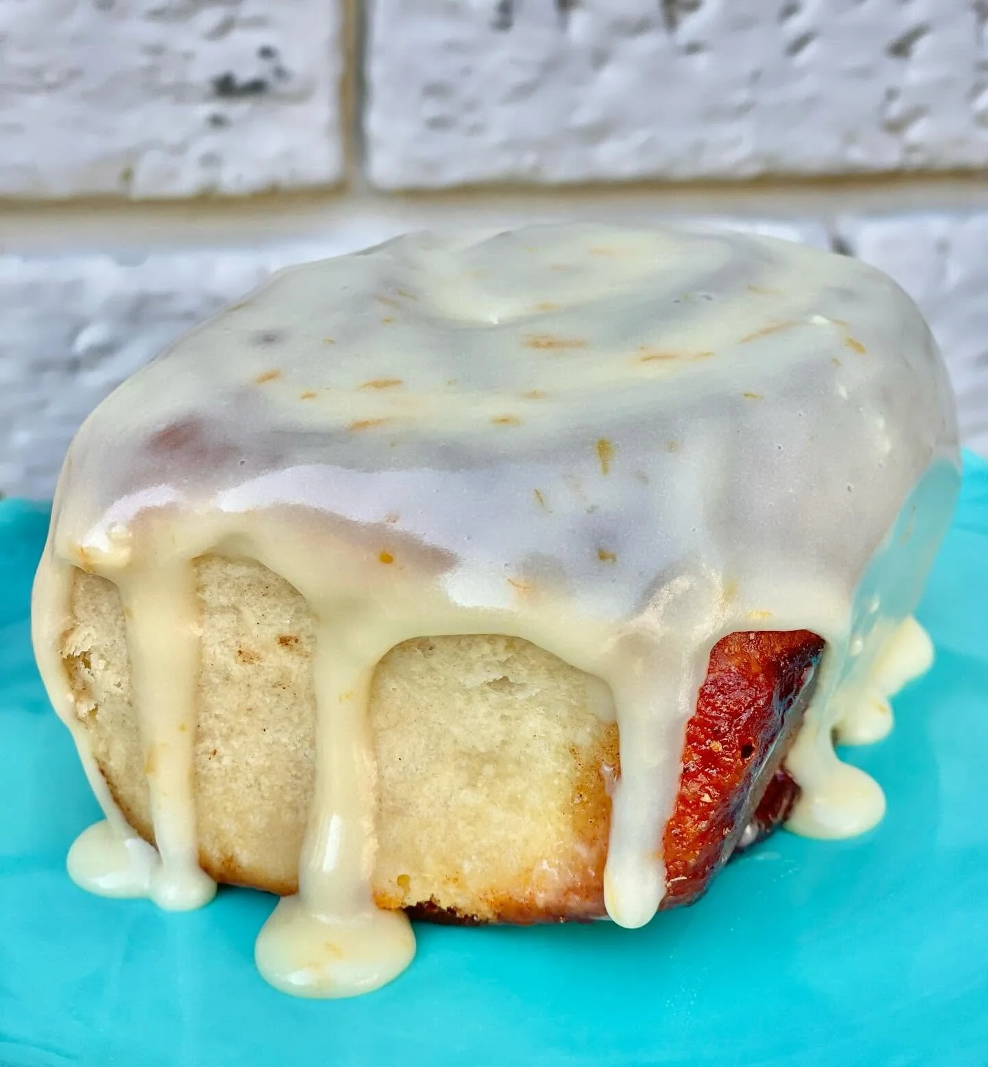 Close-up of a strawberry and vanilla cupcake with cream cheese or vanilla frosting, topped with a smooth glaze, on a light blue surface against a white brick wall background.
