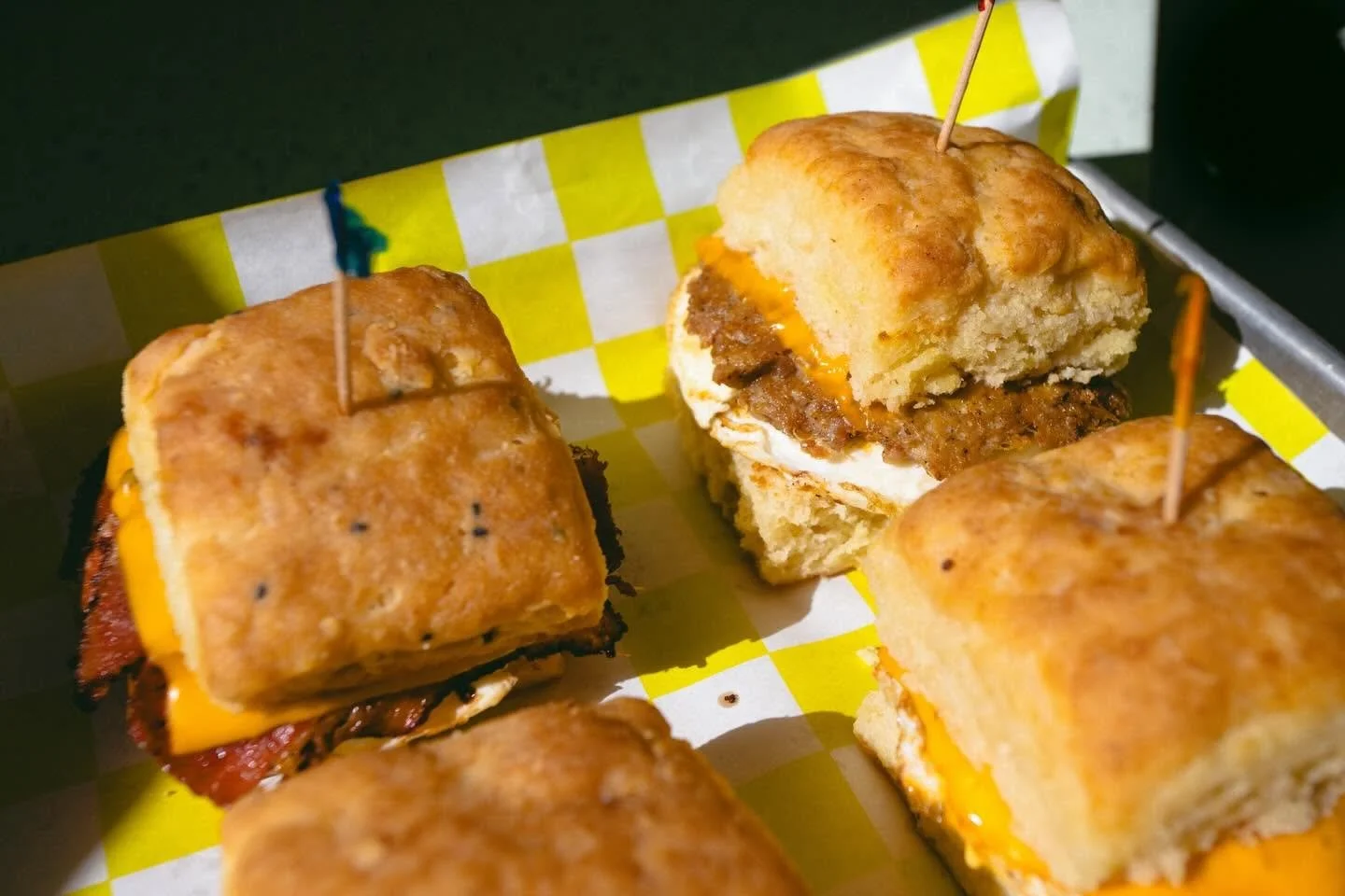 Close-up of mini cheeseburgers with biscuits on top, served on checkered paper in a metal tray.