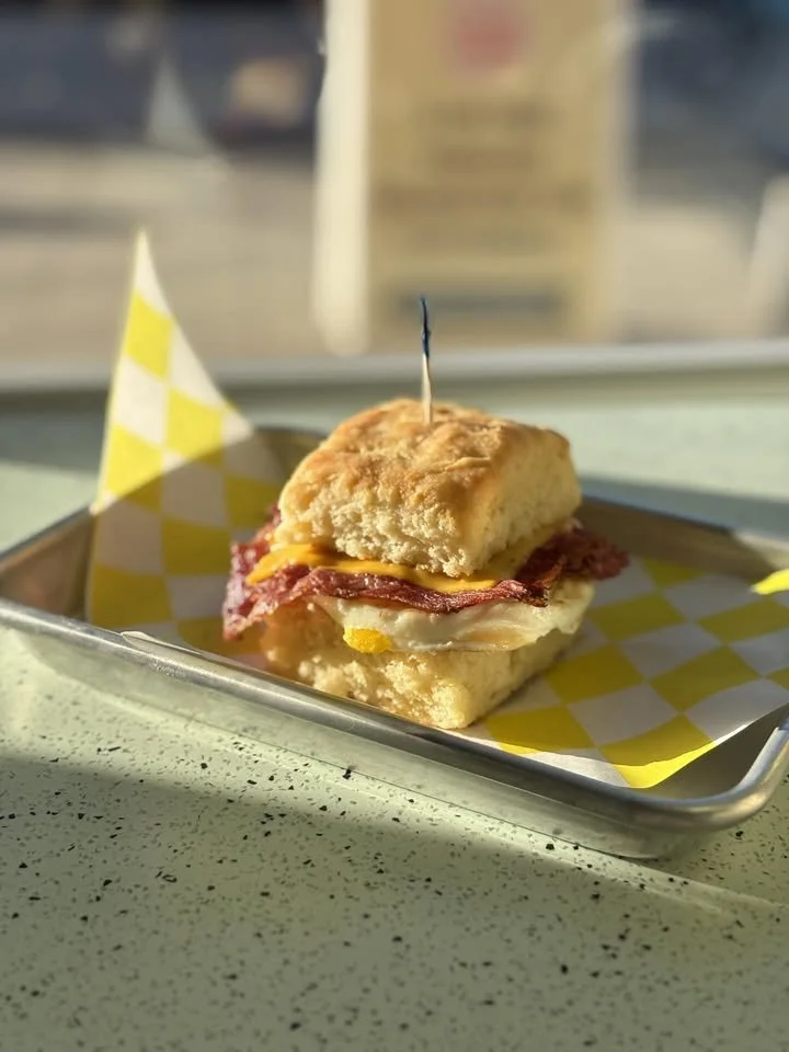 A small breakfast sandwich with biscuit, egg, bacon, and cheese on a yellow and white checkered paper in a metal tray.