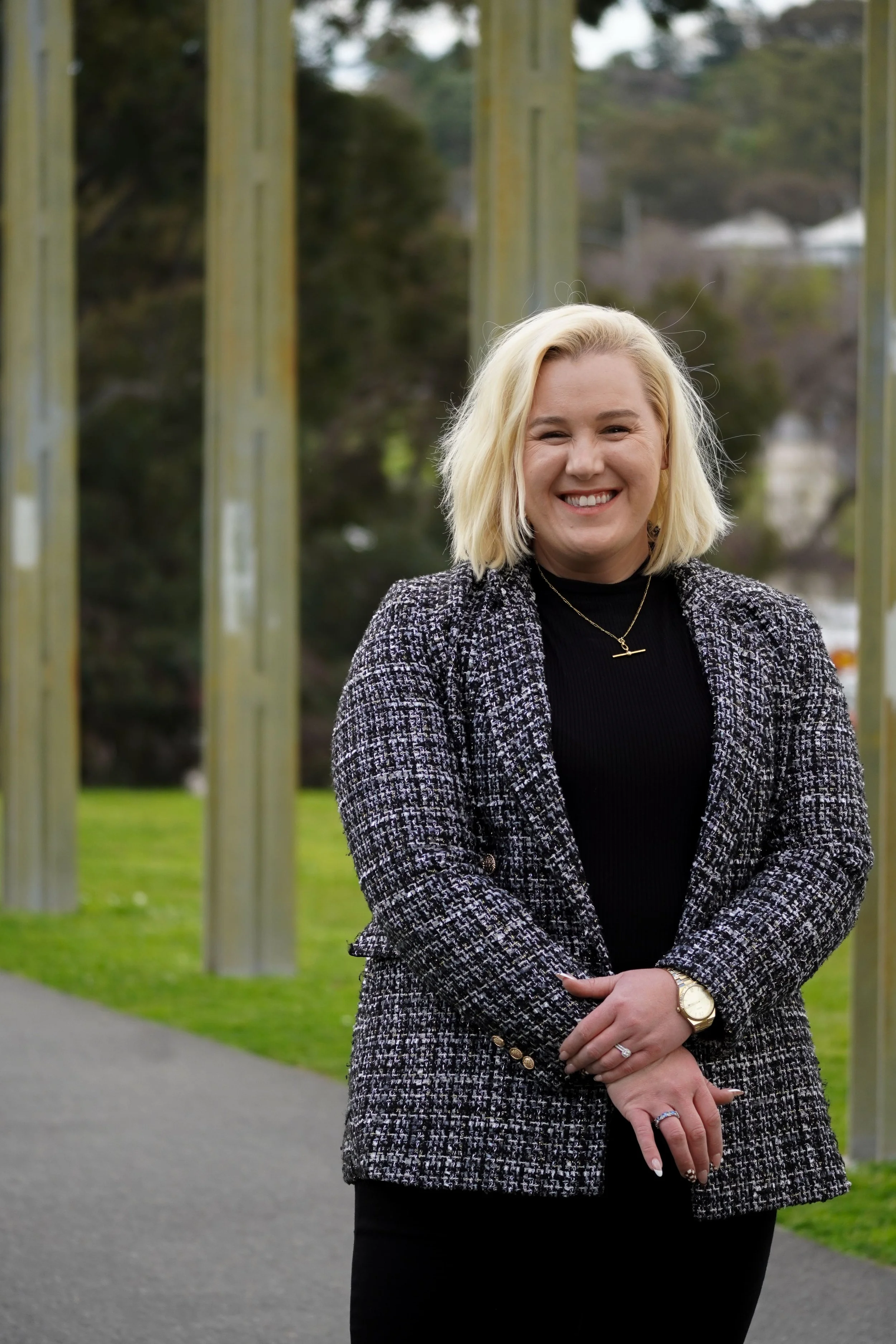 A smiling woman with blonde hair wearing a black turtleneck, a patterned black and white blazer, and black pants standing outdoors.