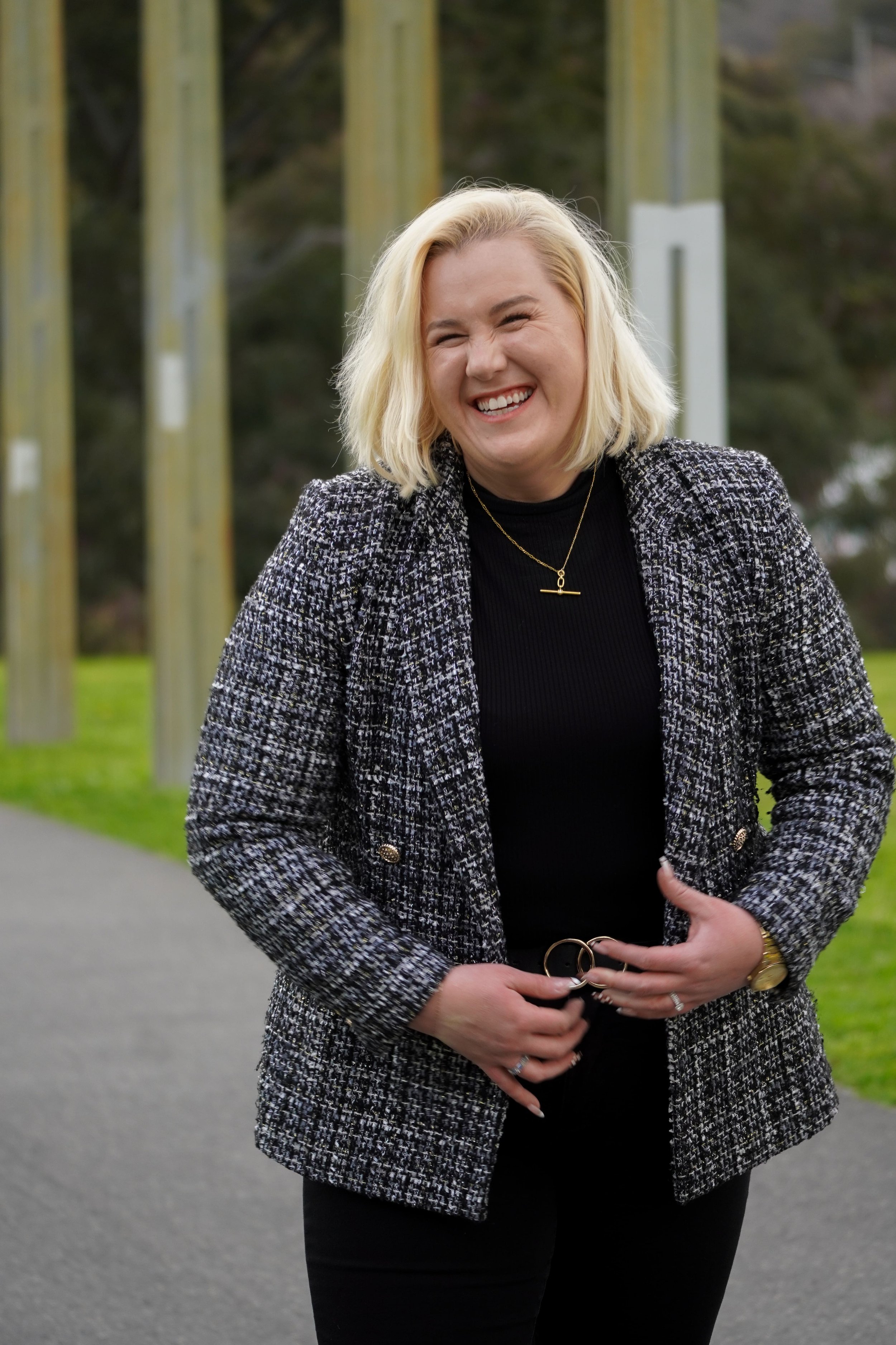 A woman with blonde hair smiling outdoors, wearing a black turtleneck, a patterned black and white blazer, and black pants.
