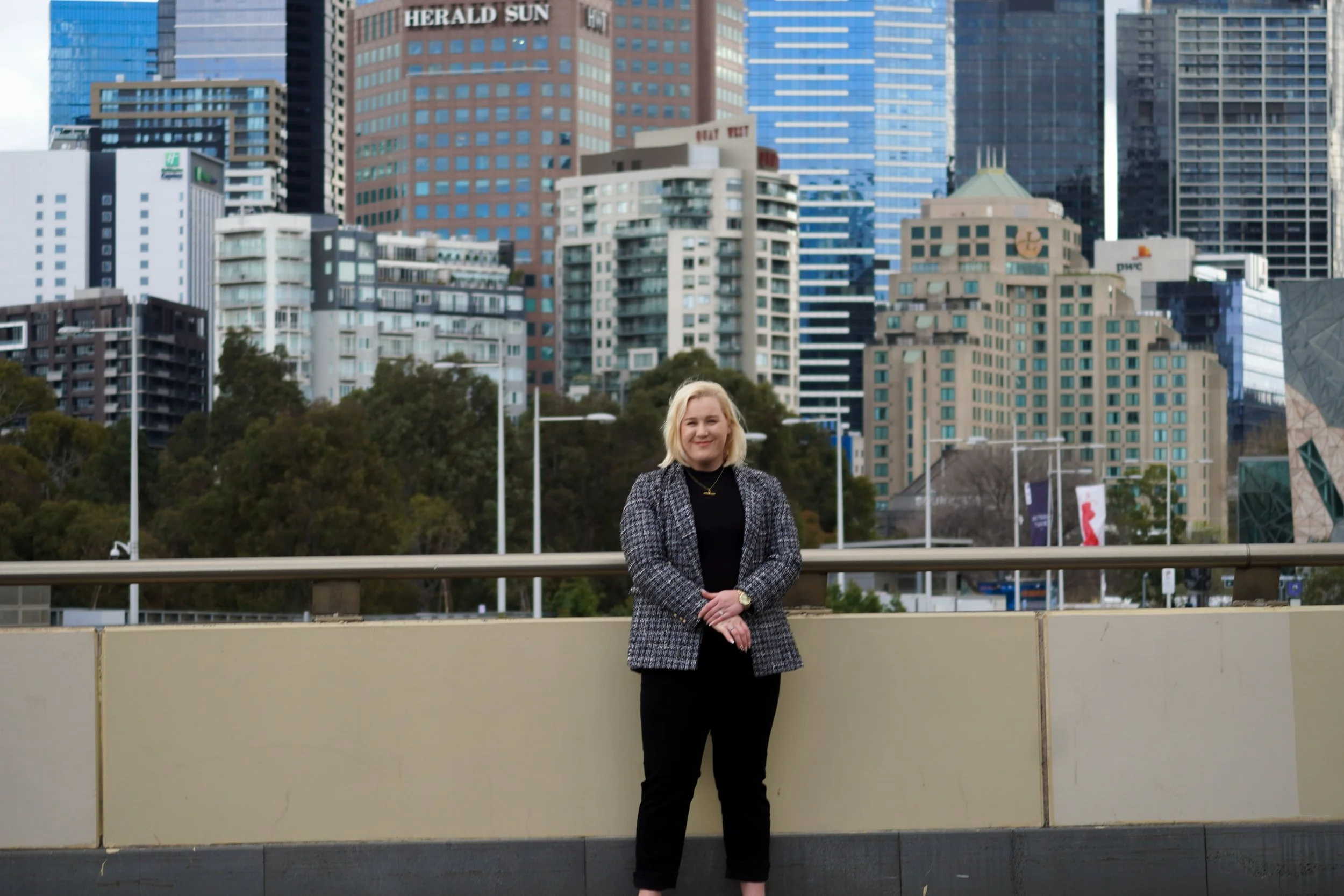 A woman with blonde hair wearing a black top and a gray checkered blazer standing outdoors in front of a city skyline with tall modern buildings and skyscrapers.